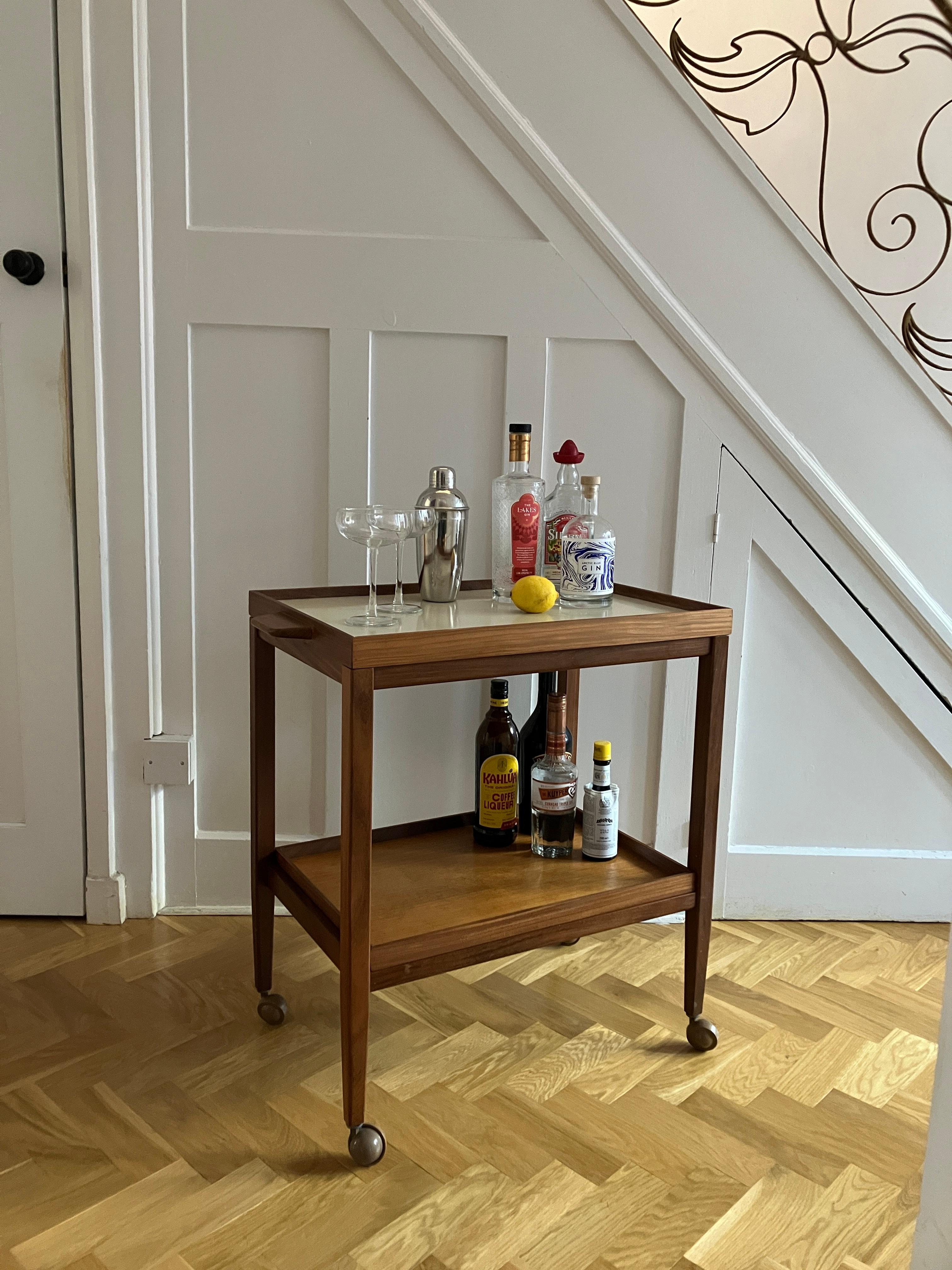 A handsome mid-century drinks trolley in solid teak. The vintage drinks trolley has two tiers with trays and both of those are detachable. The top tray is also reversible and has a cream-colored melamine cover on one side. The top tray has gorgeous