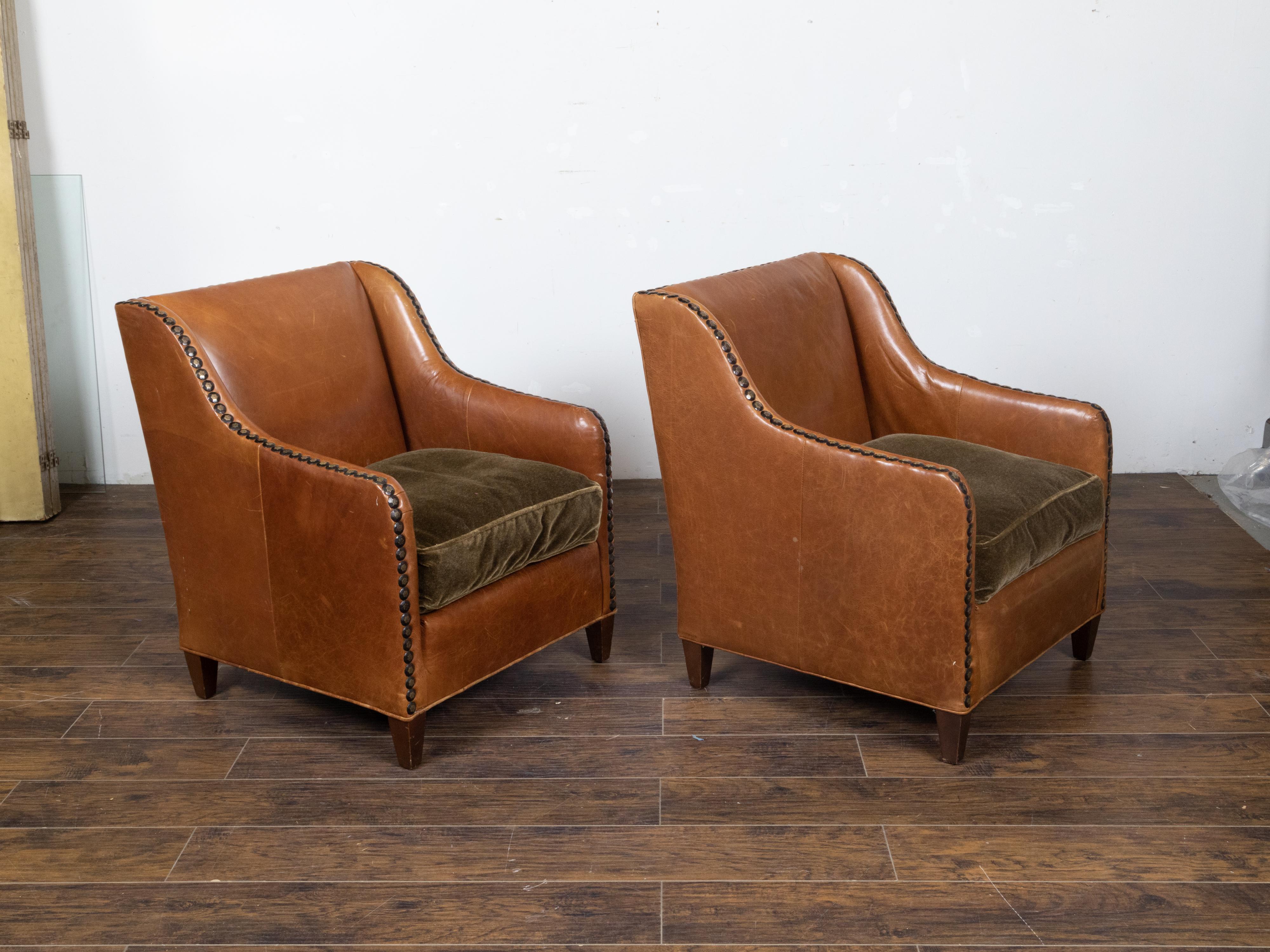 A pair of vintage English brown leather club chairs from the mid 20th century with slanted backs, velvet cushions, flattened nailhead trim and short tapered wooden feet. This pair of vintage English brown leather club chairs, hailing from the