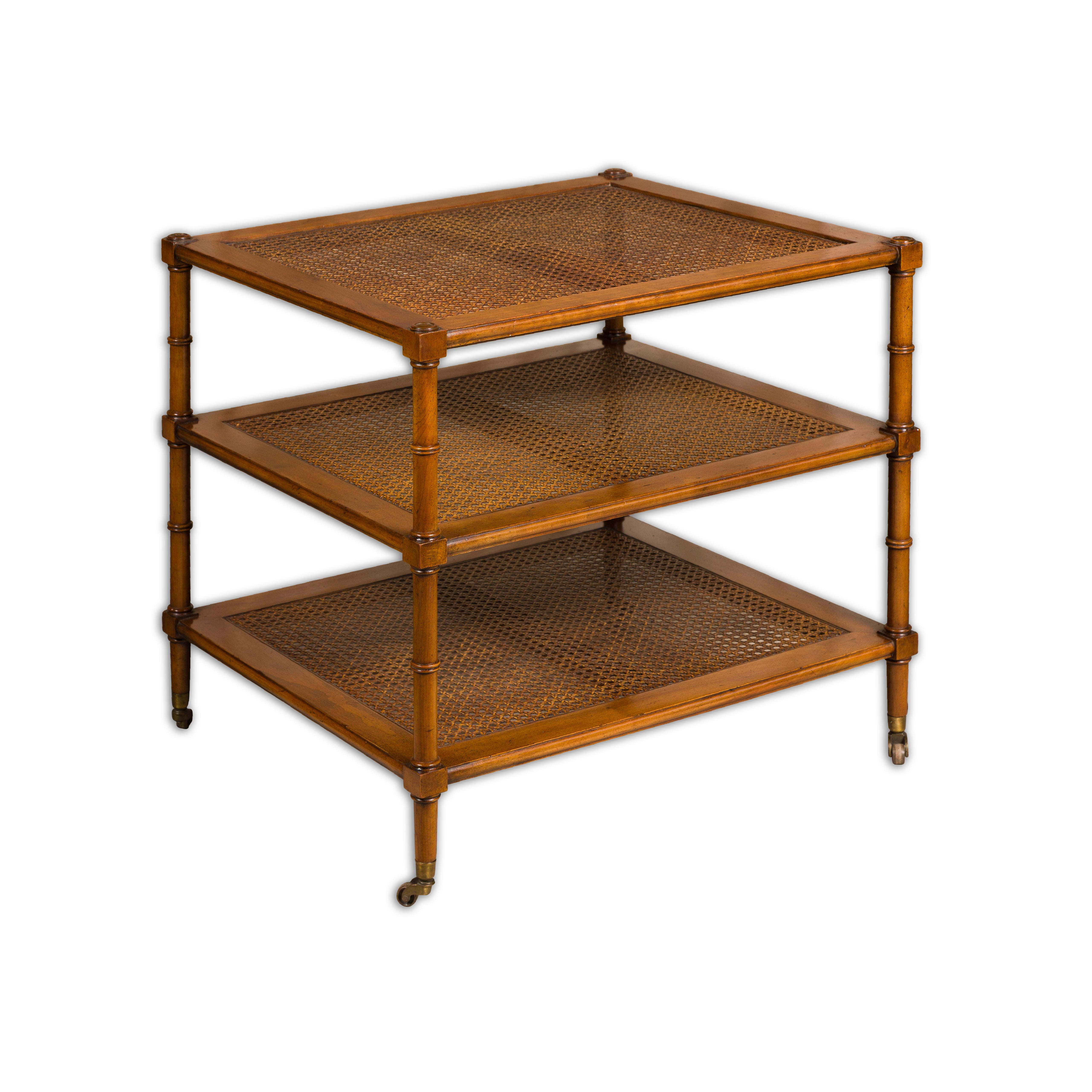 An English faux bamboo tiered table on casters with three cane shelves and casters. This English faux bamboo tiered table on casters is a delightful blend of style and functionality. Its charming design draws inspiration from the classic bamboo