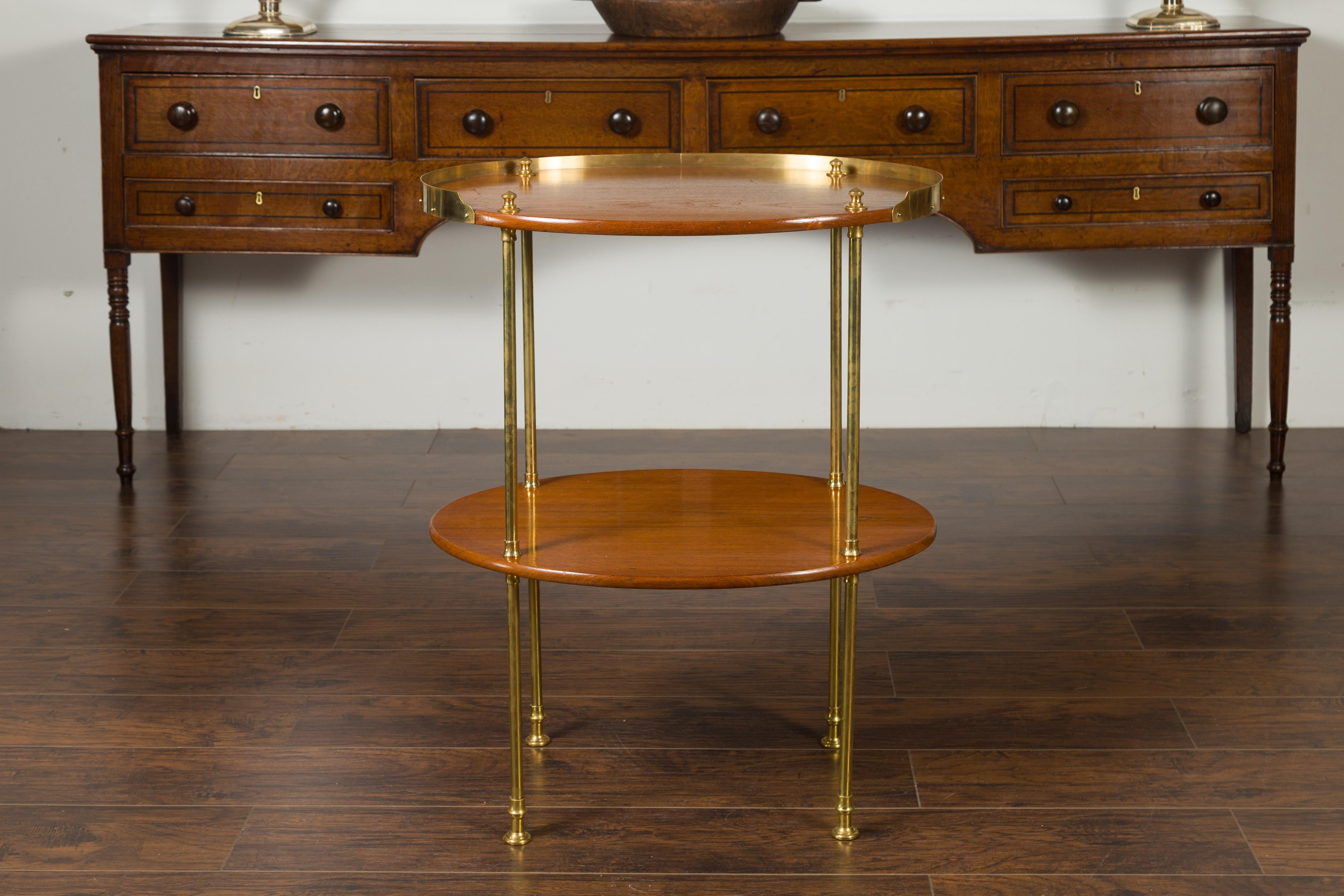 An English mahogany tiered oval table from the mid-20th century, with brass accents. Born in England during the midcentury period, this two-tiered table features an oval top surrounded by a three-quarter brass gallery, supported by brass supports