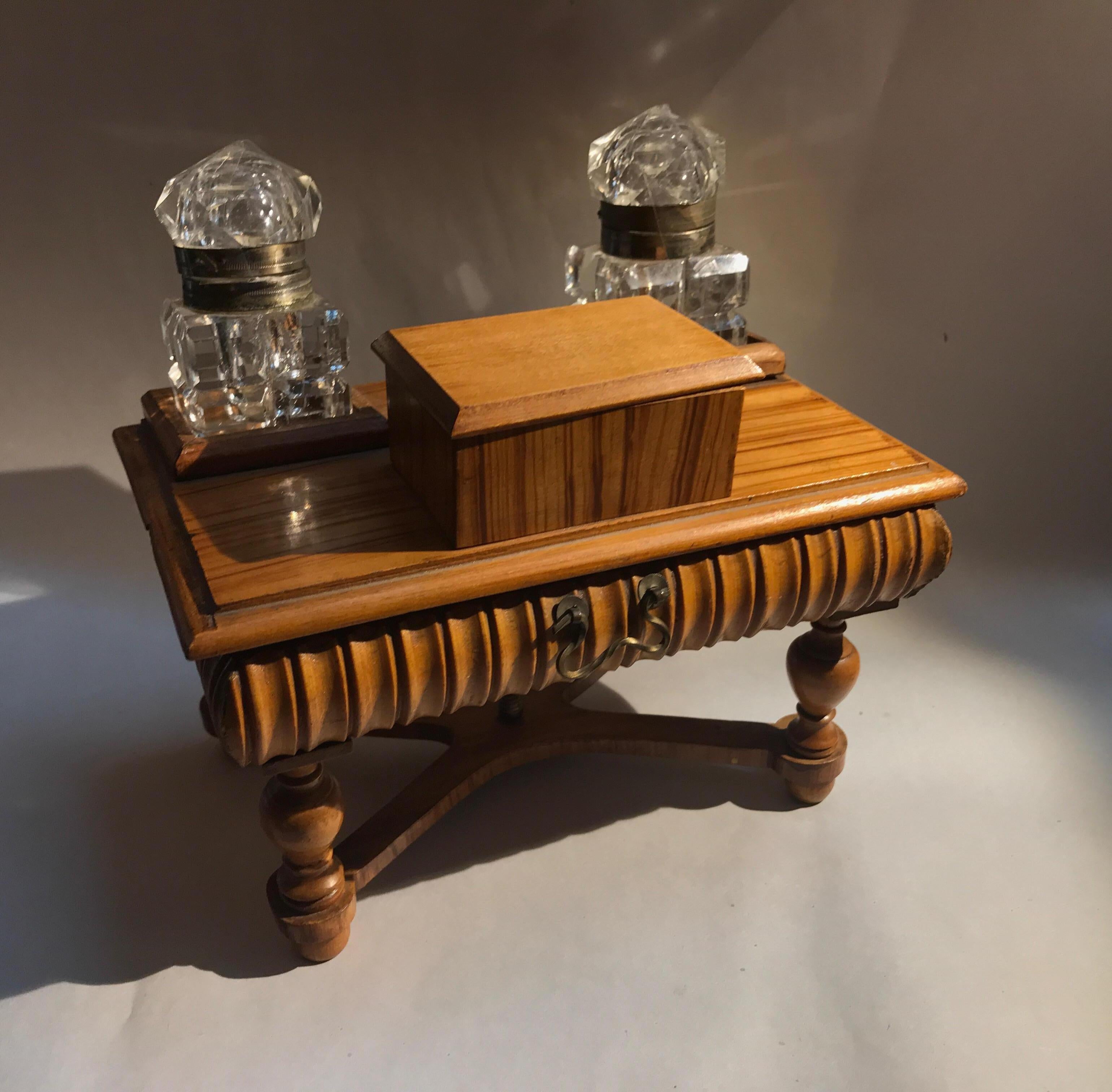 A miniature desk form inkwell and box. A double inkwell with stamp box in elm wood. The two cut-glass inkwells flank the stamp box on the top of the desk. The desk top pivots open to reveal a small storage box for pins, pen nibs and other small desk