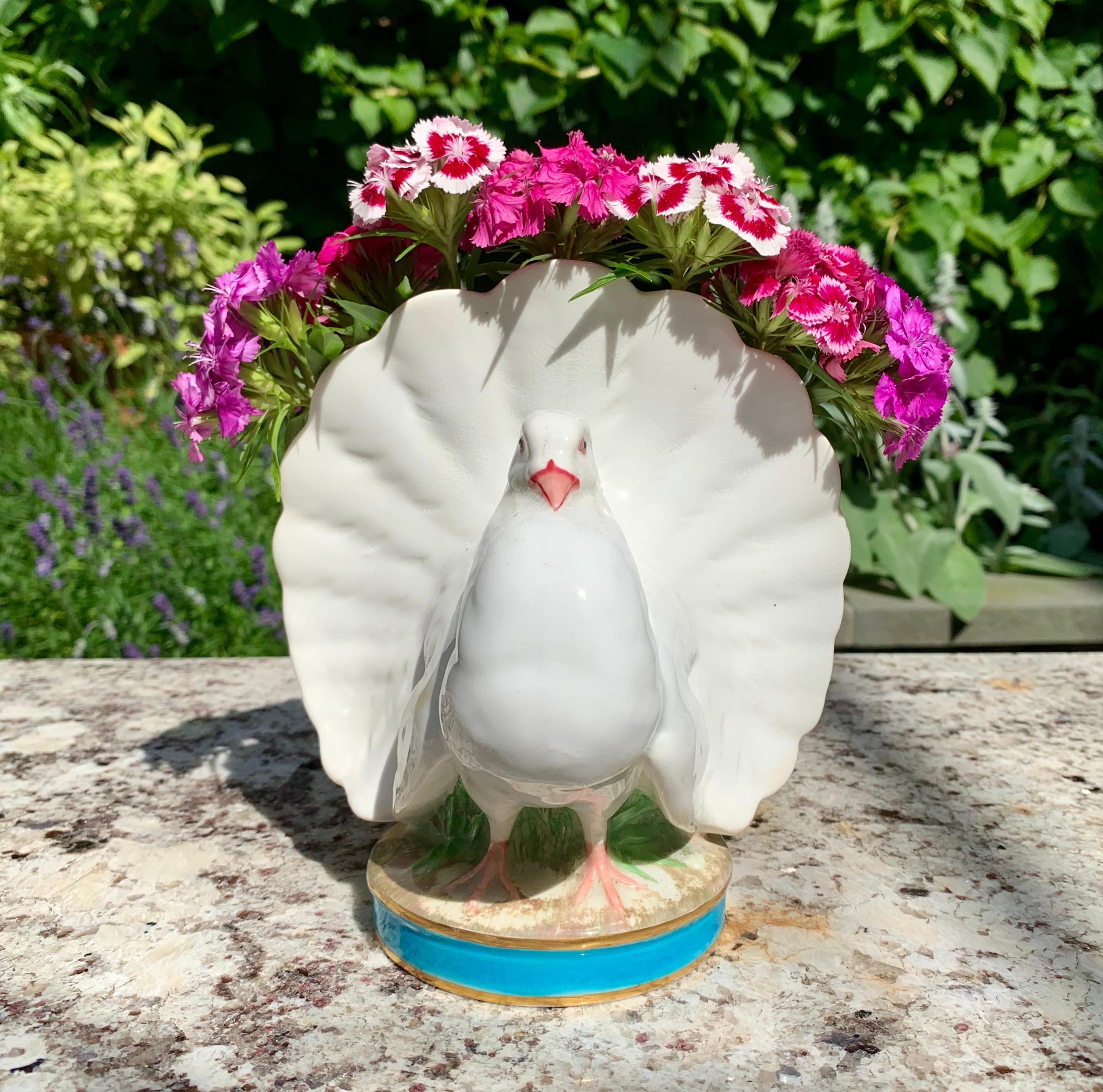 From Minton, Stoke-on-Trent, England, a porcelaneous posy vase formed as a full tailed dove or pigeon, dated 1876.

In the Rococo Revival style, the white bird sits on a round grassy base banded in turquoise and gold. Her flared tail feathers