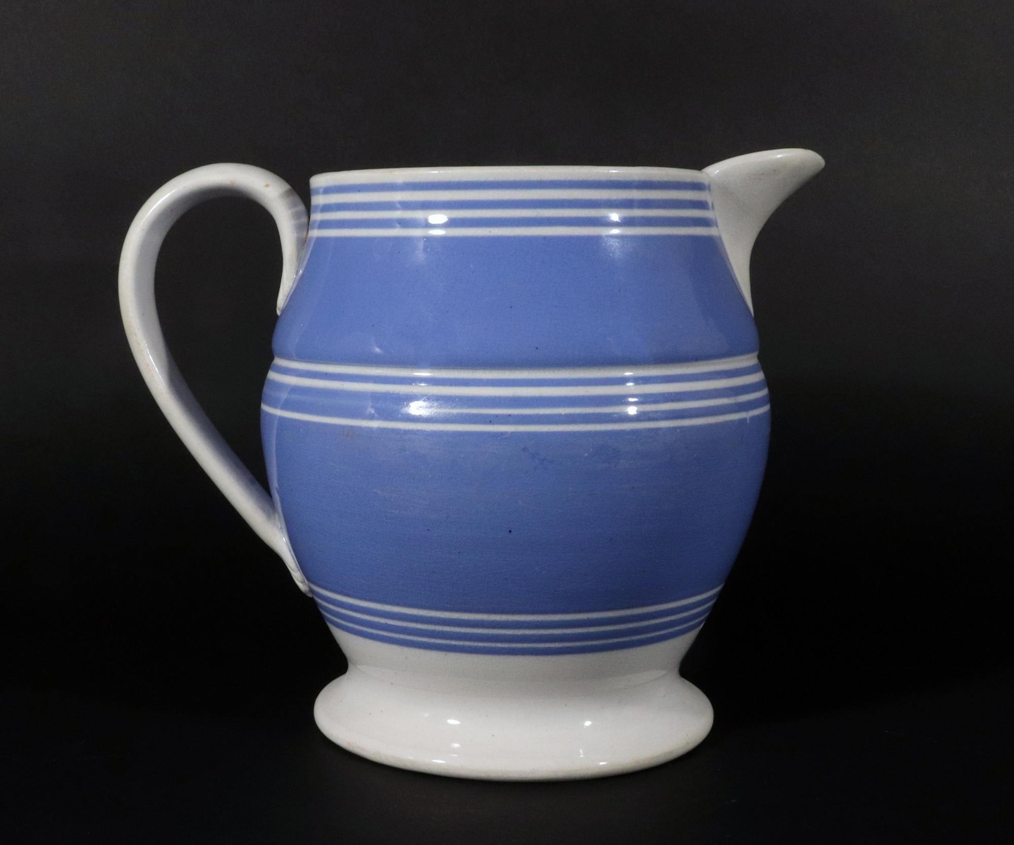 English Mocha-decorated Pottery Blue Slip Jug with White Bands,
Circa 1820

This slip-decorated pearlware jug features a vibrant blue slip ground adorned with three distinct groupings of four thin white bands. The bands encircle the body of the jug,