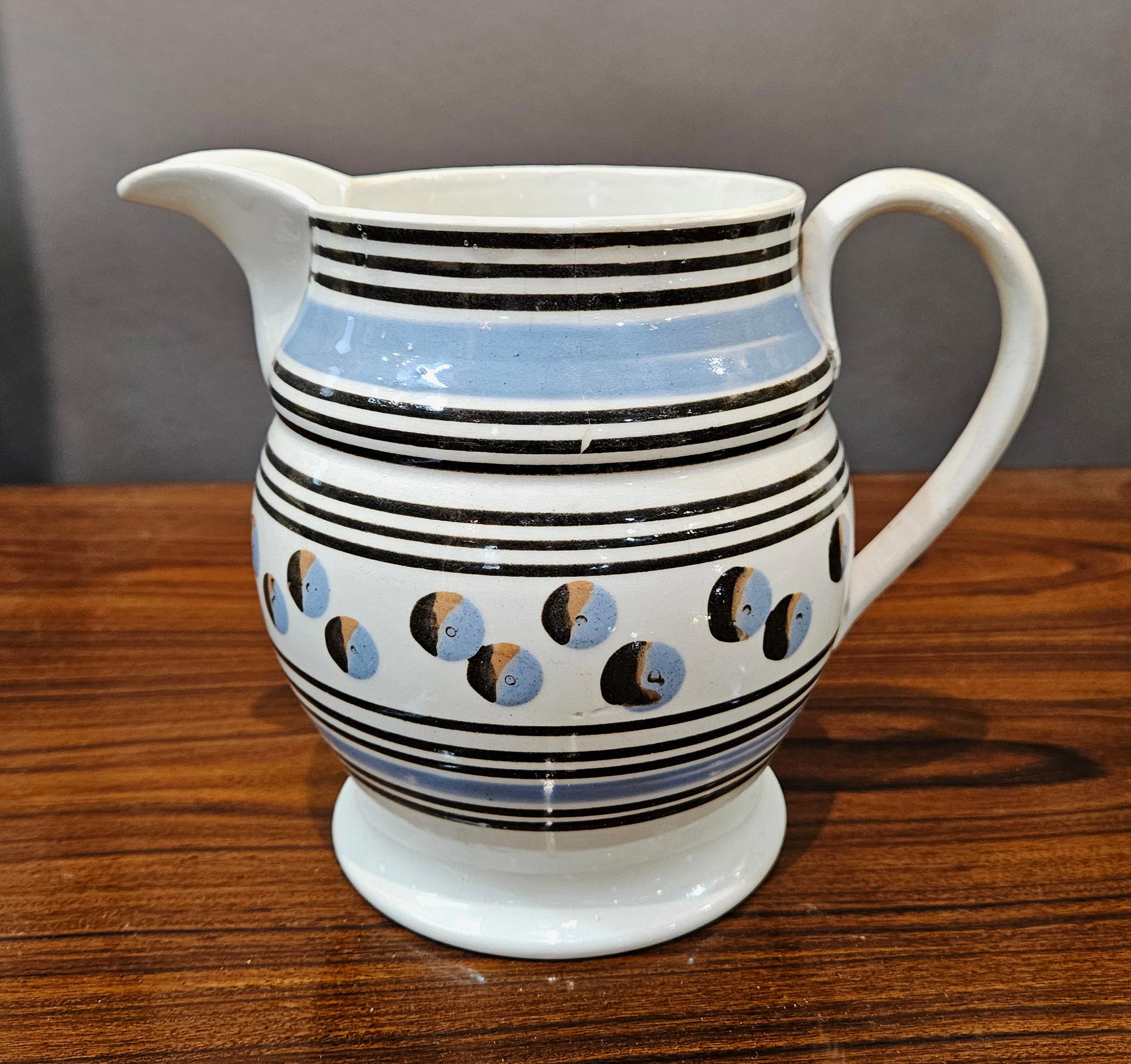 English Mocha Pottery Pearlware Cat's Eye Krug,
CIRCA 1800-1820

Die Mokka-Kanne aus Perlmutt hat ein Band aus dreifarbigen Katzenaugen-Motiven  in Braun, Orange und Blau mit einer Reihe von Bändern oben und unten in Braun und Hellblau. Der