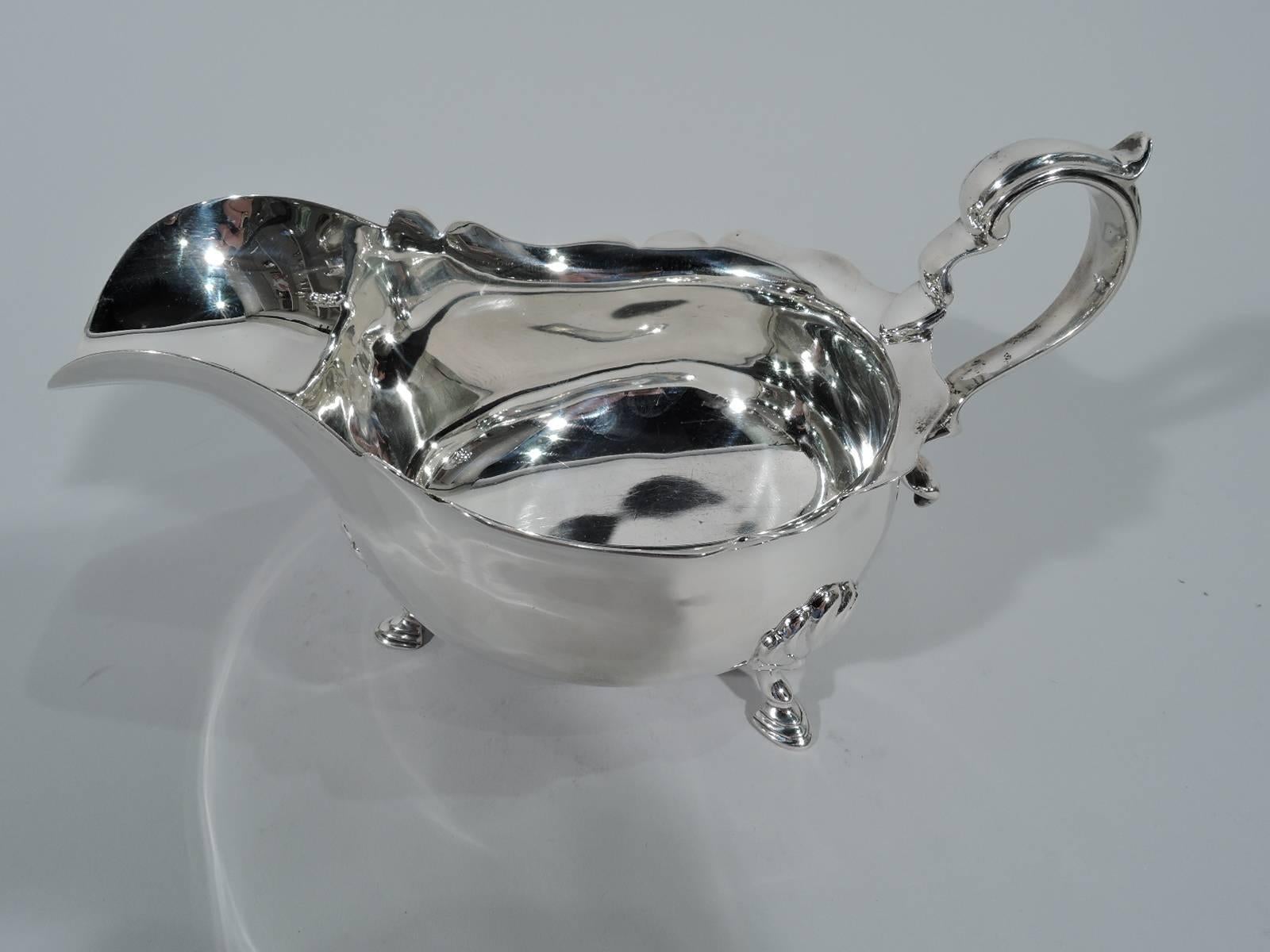 George VI sterling silver gravy boat. Made in London in 1939. Wide-bodied bowl with scalloped rim, helmet mouth, capped-scroll handle, and 3 shell-mounted hoof supports. Traditional Georgian – still vital after more than two centuries. Fully marked