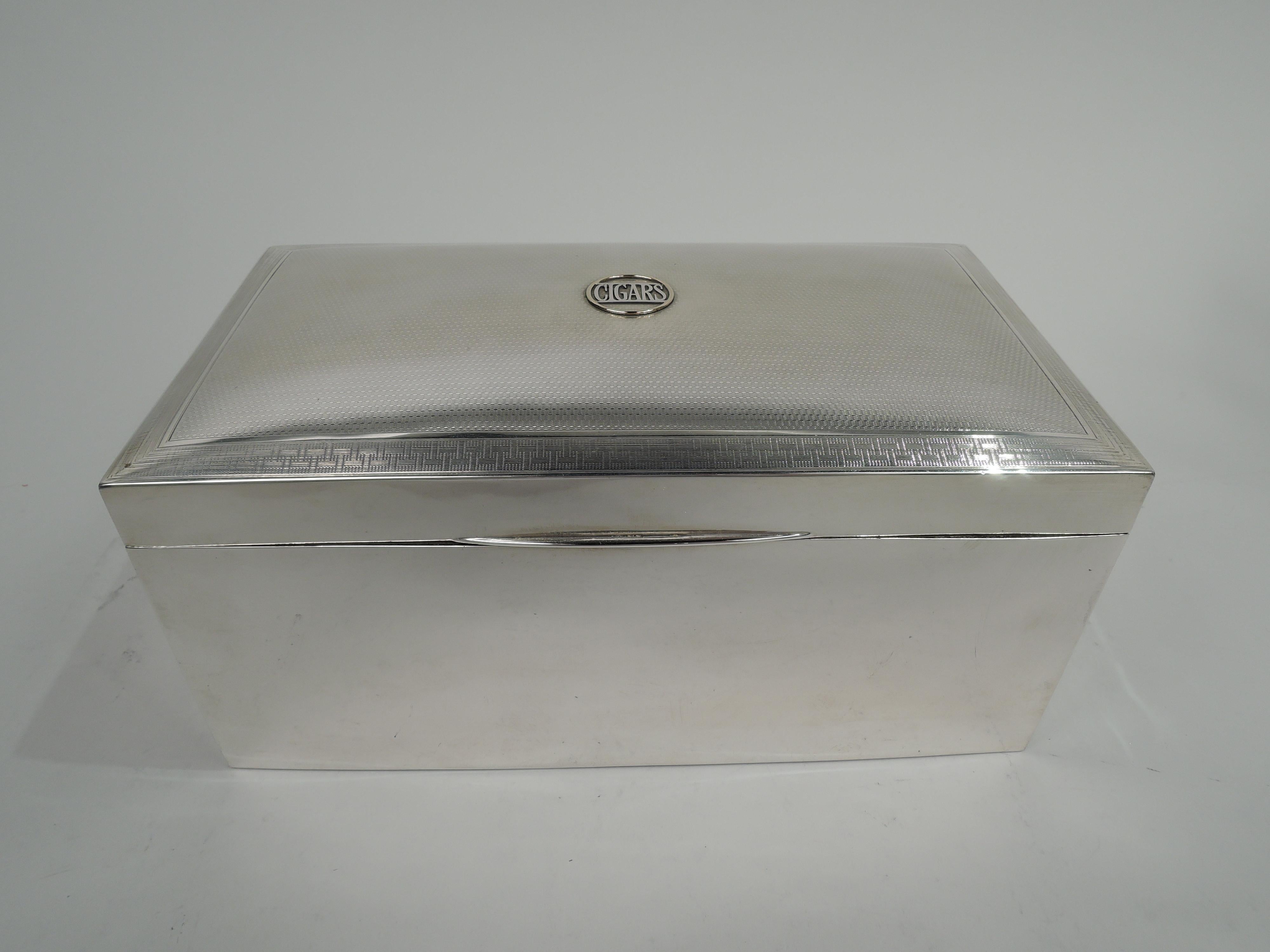 English Modern sterling silver cigar box, 1919. Rectangular with plain straight sides. Cover hinged with tapering tab; top gently curved with engine-turned wave ornament bordered by fretwork, and centrally mounted gold ring inset with word “CIGARS”.