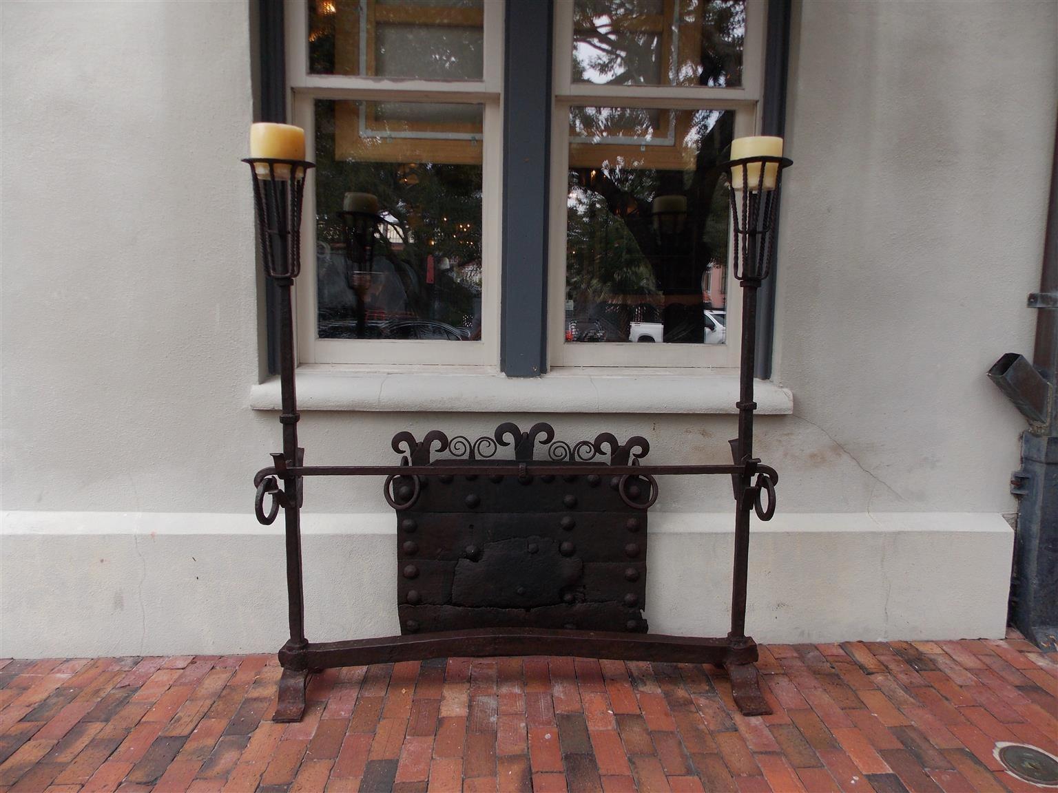 English monumental wrought iron fire guard with decorative scrolled medallion fire back and flanking wax candle holders, Mid-18th century.

Measurements
Andirons 66.75 tall, 65 wide, 13.5 deep
Fire back 33.25 tall, 38 wide.