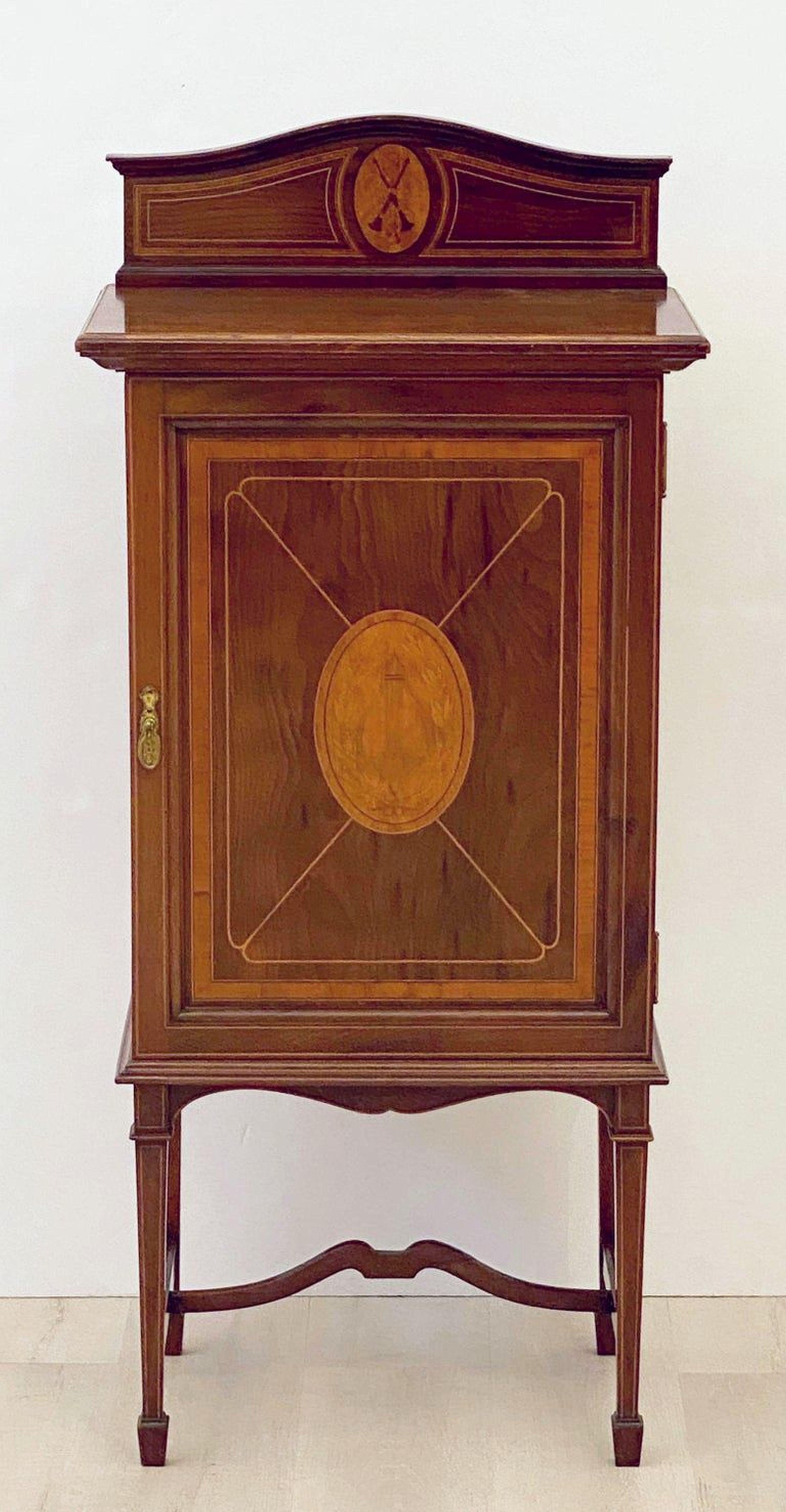 A fine English music cabinet or cupboard of inlaid mahogany from the Edwardian era, featuring a moulded top with shaped back panel and inlaid design of two wind instruments in a center cartouche, over a cupboard with paneled door. The door with