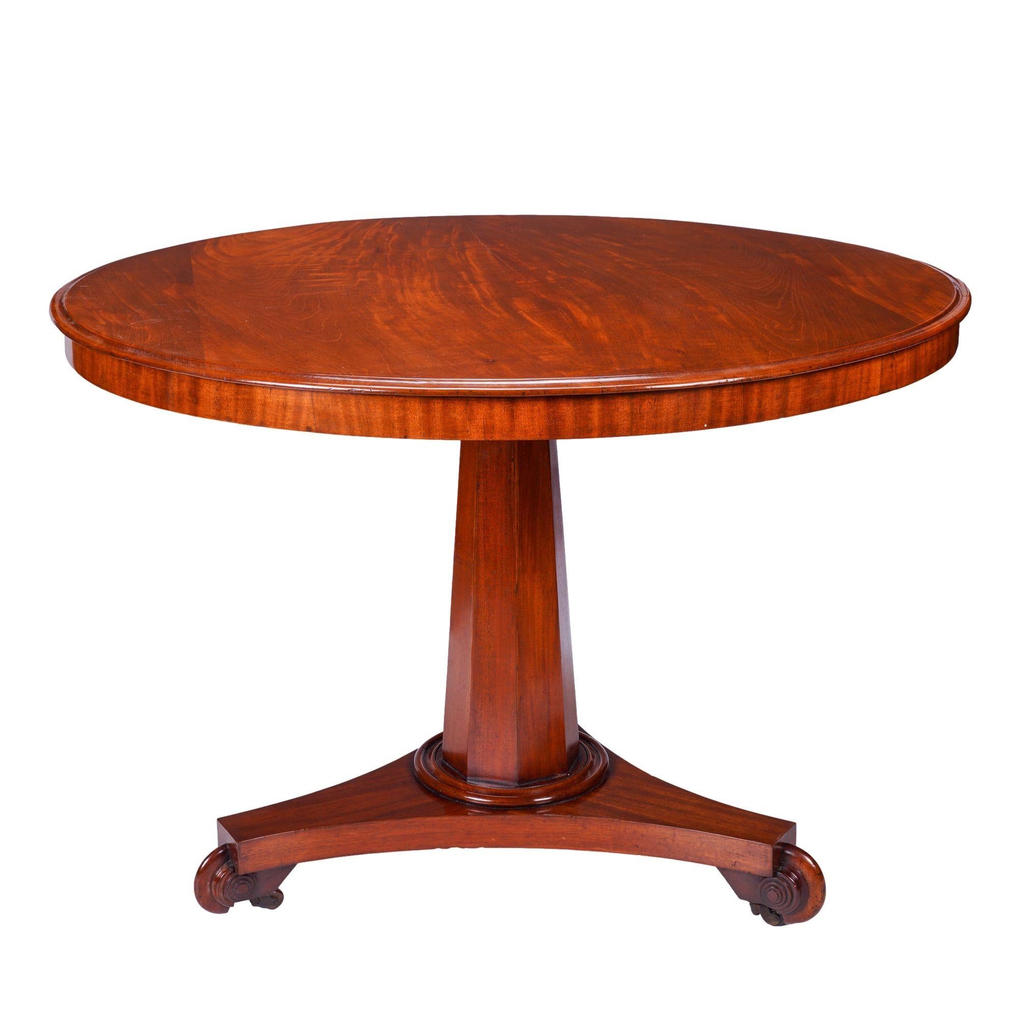 English Neoclassic figured mahogany circular tilt top table with ogee edge above a shallow cross banded apron. The table top is supported by an octagonal tapered columnar pedestal on a shallow ring turned molding and mounted on concave trefoil