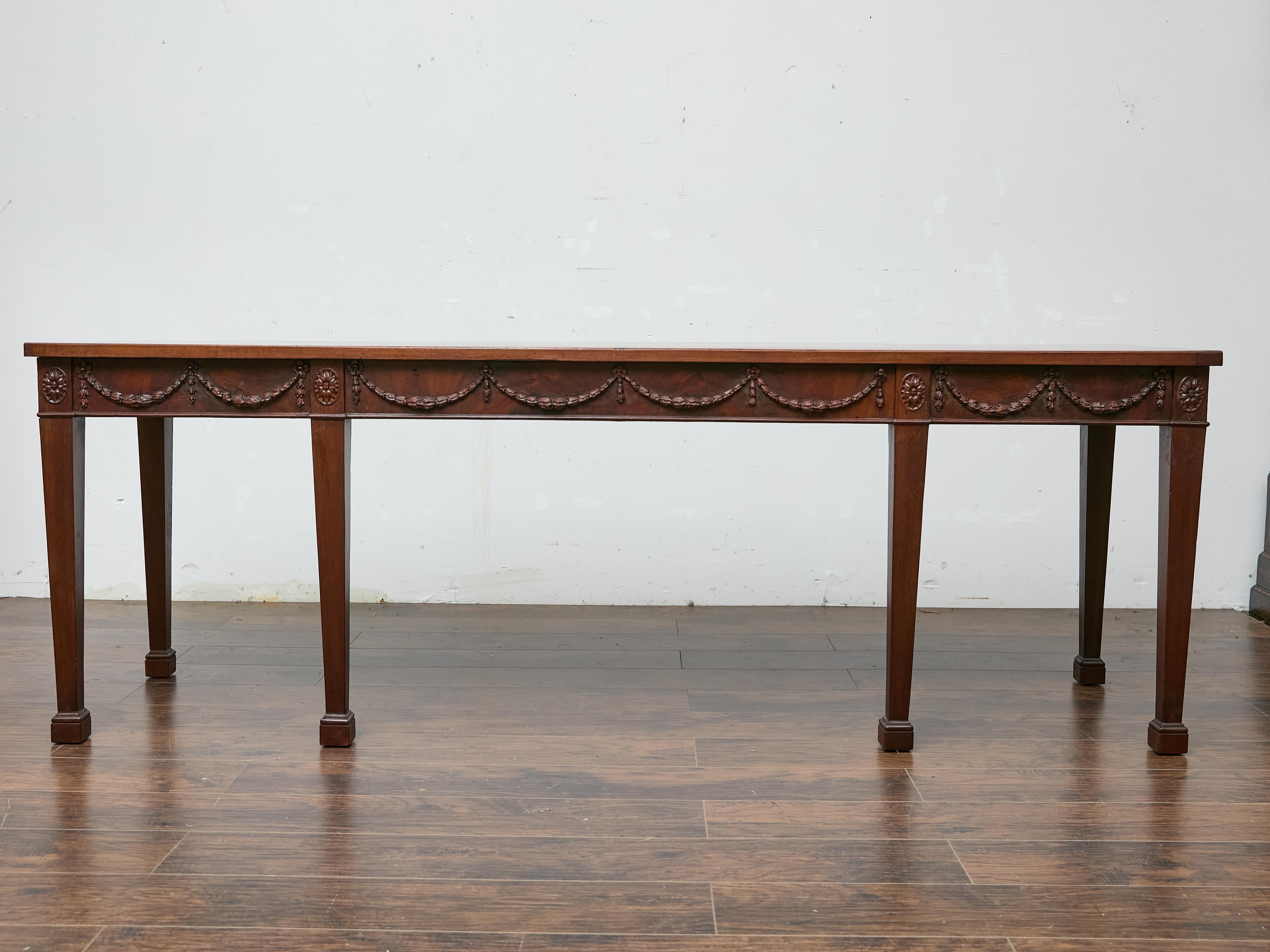Crafted in the 19th century, this English Neoclassical style console table is built from rich, figured mahogany and stands on six tapered legs with block feet. The long frieze is carved in shallow relief with swags and rosettes, an elegant