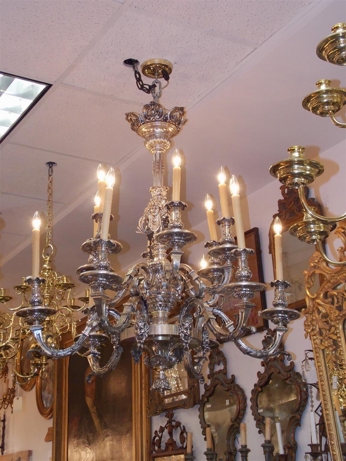 English silver over bronze figural chandelier with decorative floral canopy, centered fluted berry and mask column, twelve fluted scrolled arms with acanthus foliage, and resting on a acanthus shell circular base with flanking mythological figures.