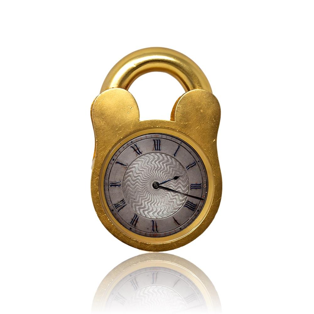 English novelty desk clock timepiece in the form of a padlock. The clock set within the body of the padlock shape with silvered dial and roman numeral around the perimeter. The clock fitted with a hinged stand to the rear with keyhole wind and time
