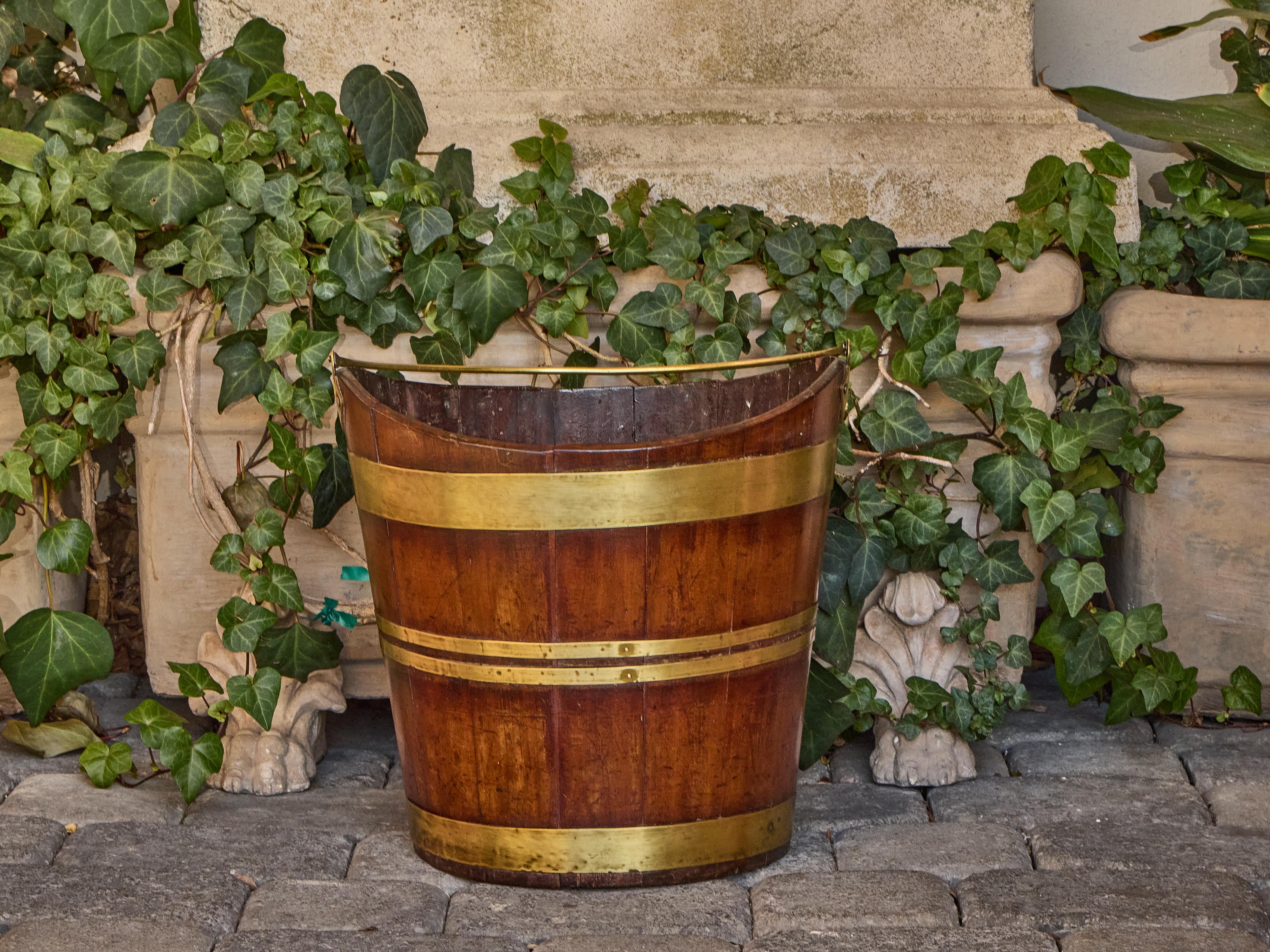 This English oak and brass bucket from the 19th century combines practical construction with sculptural form. Built from vertical oak staves in the traditional coopered manner, the bucket is reinforced with four solid brass bands, each carefully