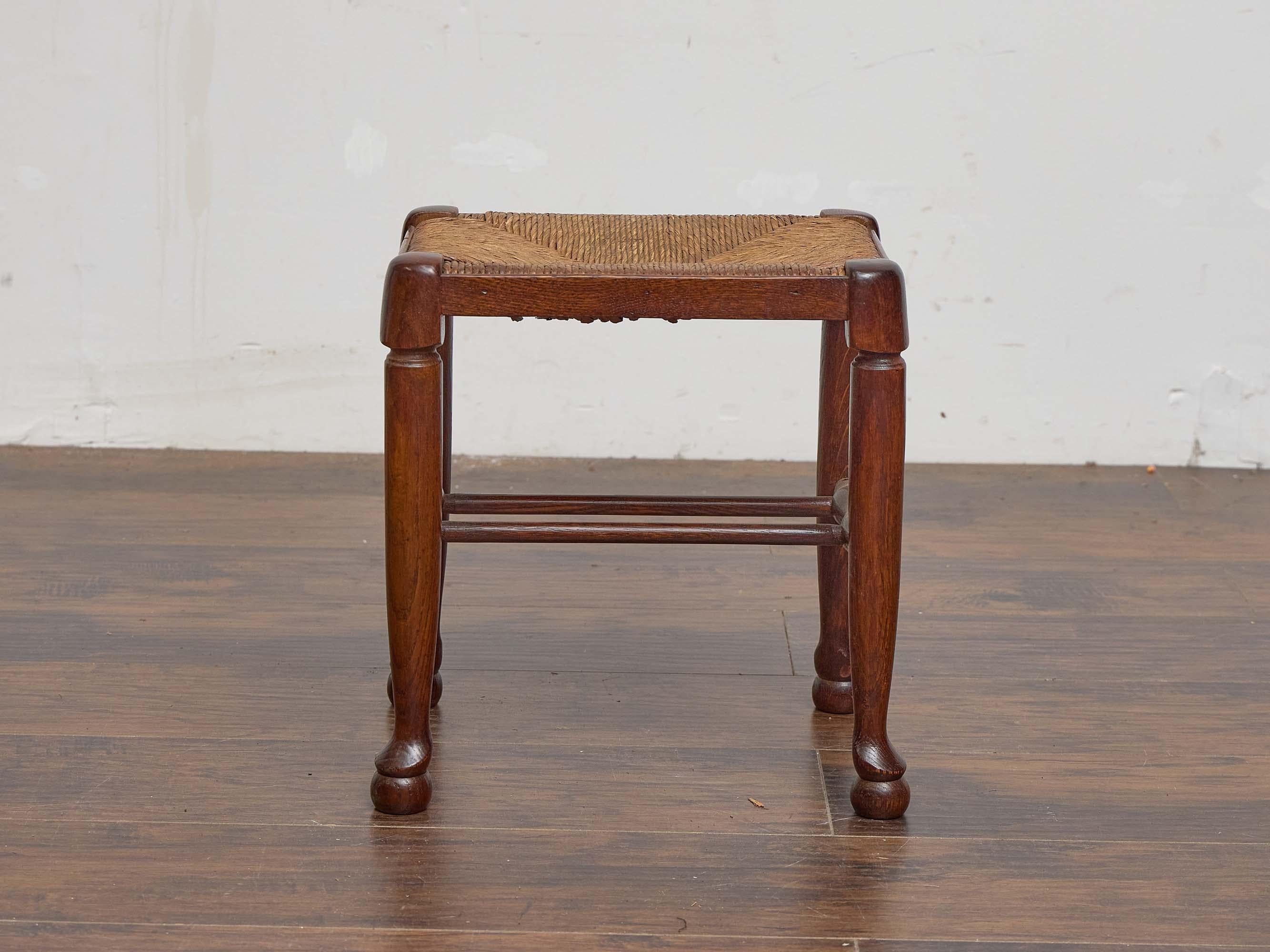 Fabriqué en Angleterre vers 1900, ce charmant tabouret en chêne est doté d'une assise en jonc tressé à la main et d'une structure subtilement sculpturale avec une menuiserie traditionnelle. L'assise carrée présente un motif classique de jonc en