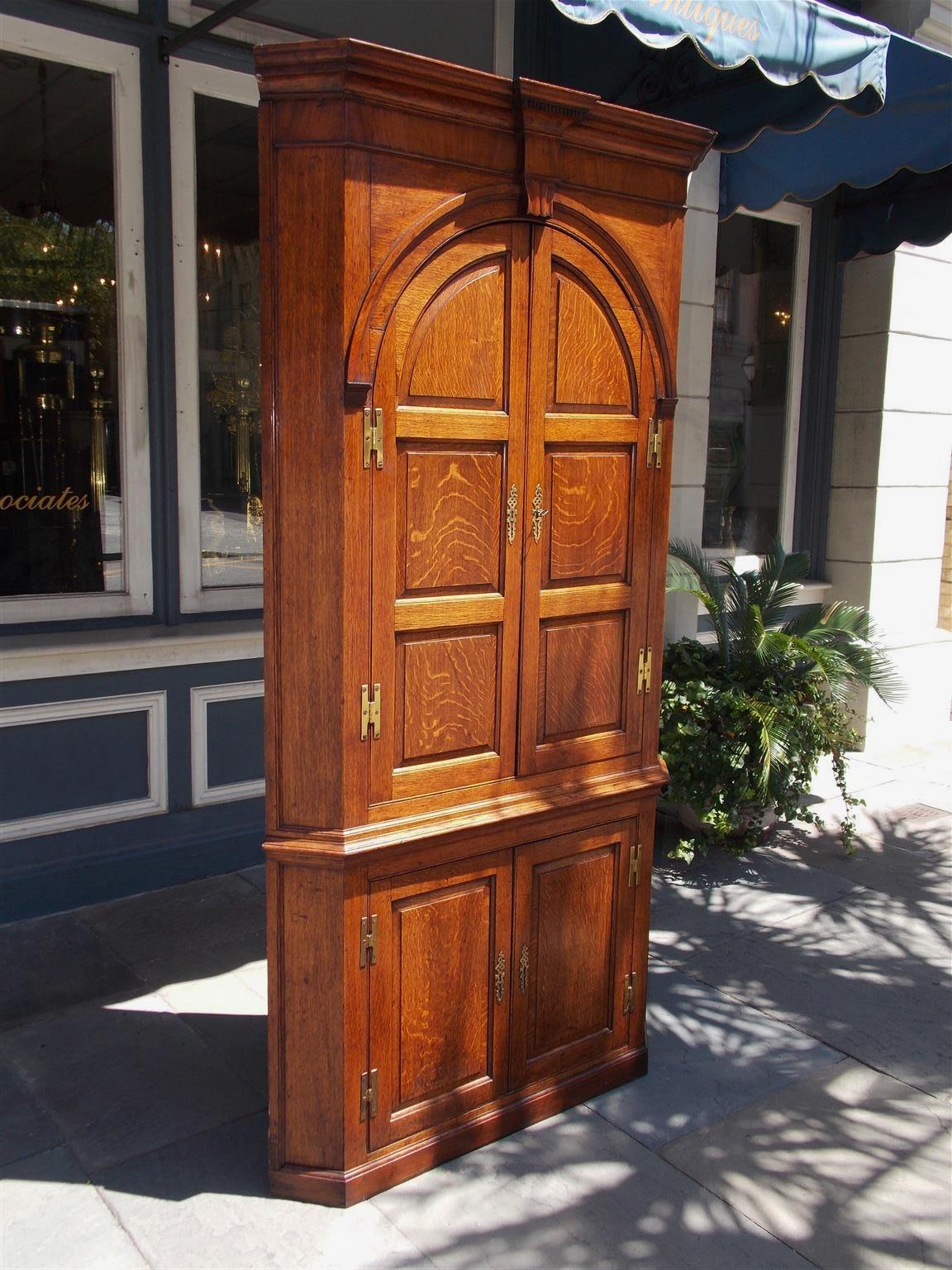 English oak blind door corner cabinet with a carved molded edge cornice, centered dental molding with gouge work, upper case with flanking H-hinged arched paneled doors revealing three butterfly shelves and three pigeon drawers, lower case with