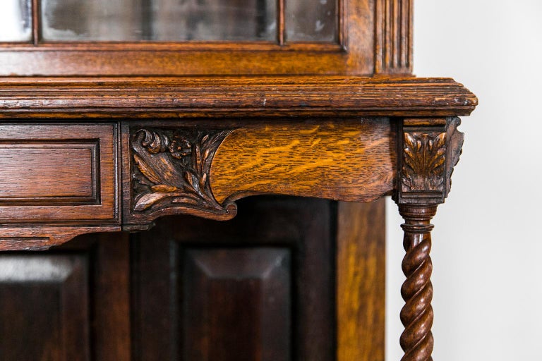 The top of this bookcase/display cabinet has a broken arch pediment with a stylized egg and dart molding carved in high relief. The platform shelf is inlaid with oak paneling and rests on barley twist front legs. The stretcher connects to a triple