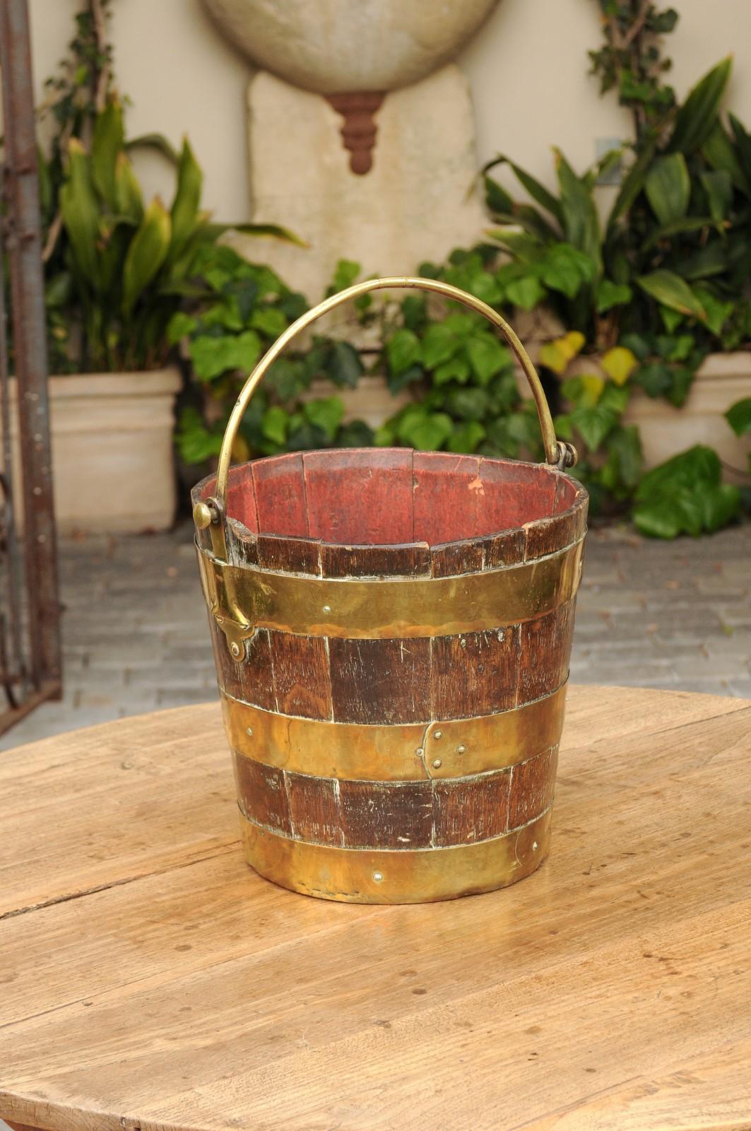 An English oak bucket from the late 19th century, with brass handle and braces. Born in England during the last quarter of the 19th century, this charming oak bucket features vertical slats reinforced by the presence of horizontal brass braces,