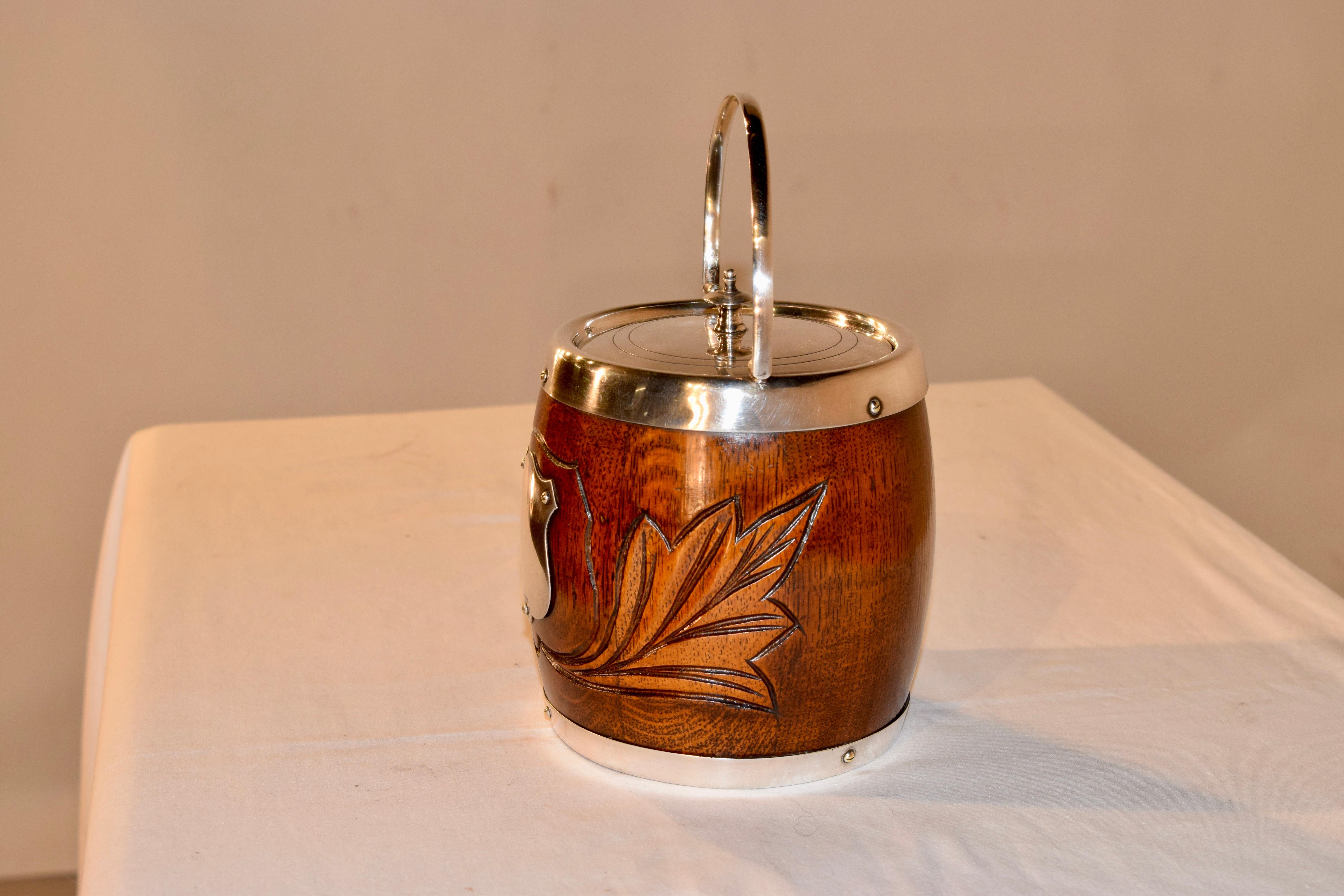 English oak biscuit barrel with silver plated lid, shield on the front and banding. It retains the original porcelain liner inside. This barrel has lovely hand-carving with a central shield and leaves to either side.
 