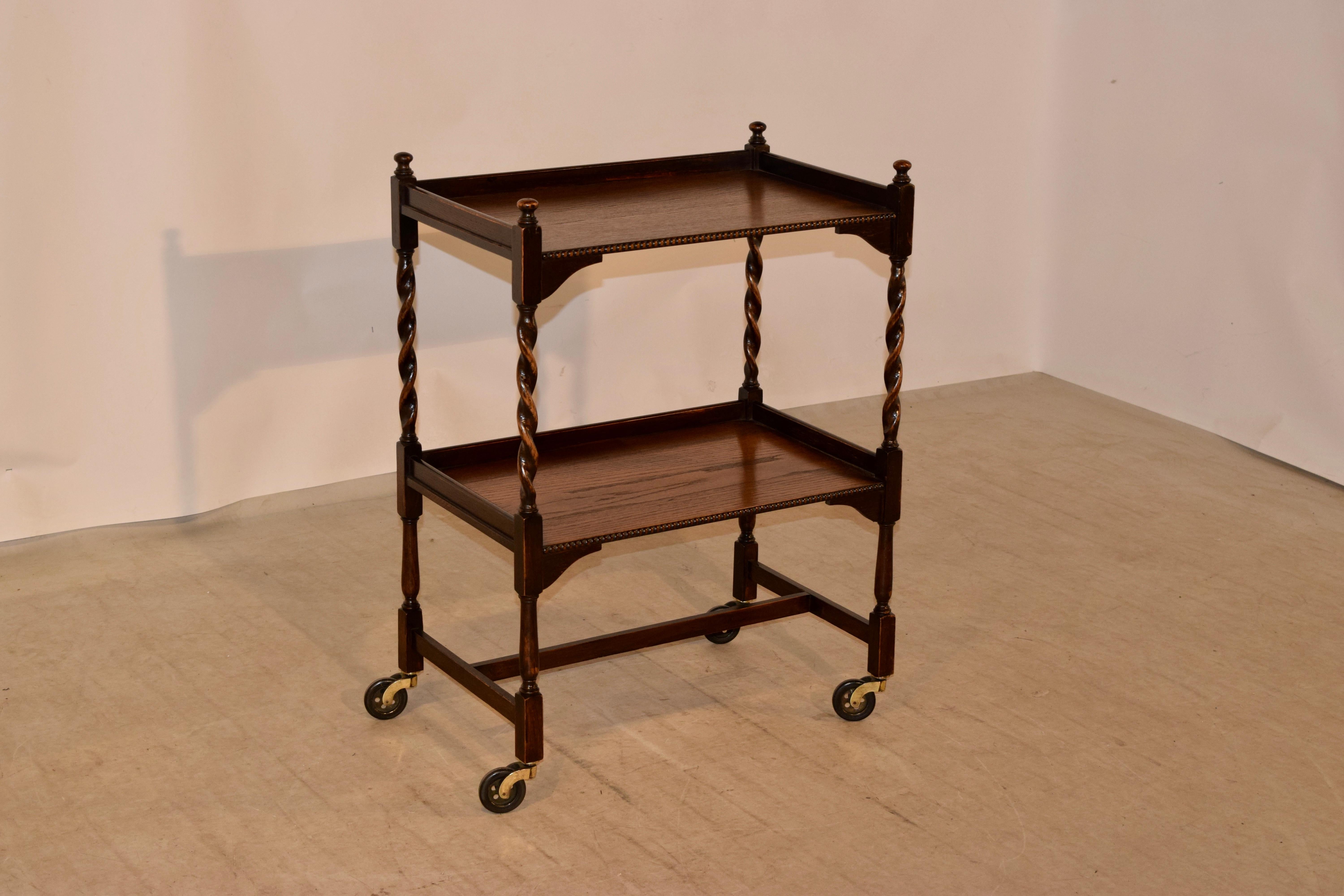 English oak drinks cart, circa 1900 with finials decorating the top following down to two shelves, both with galleries and beaded edges along the fronts. The shelves are separated by hand-turned barley twist shelf supports. The legs are hand-turned