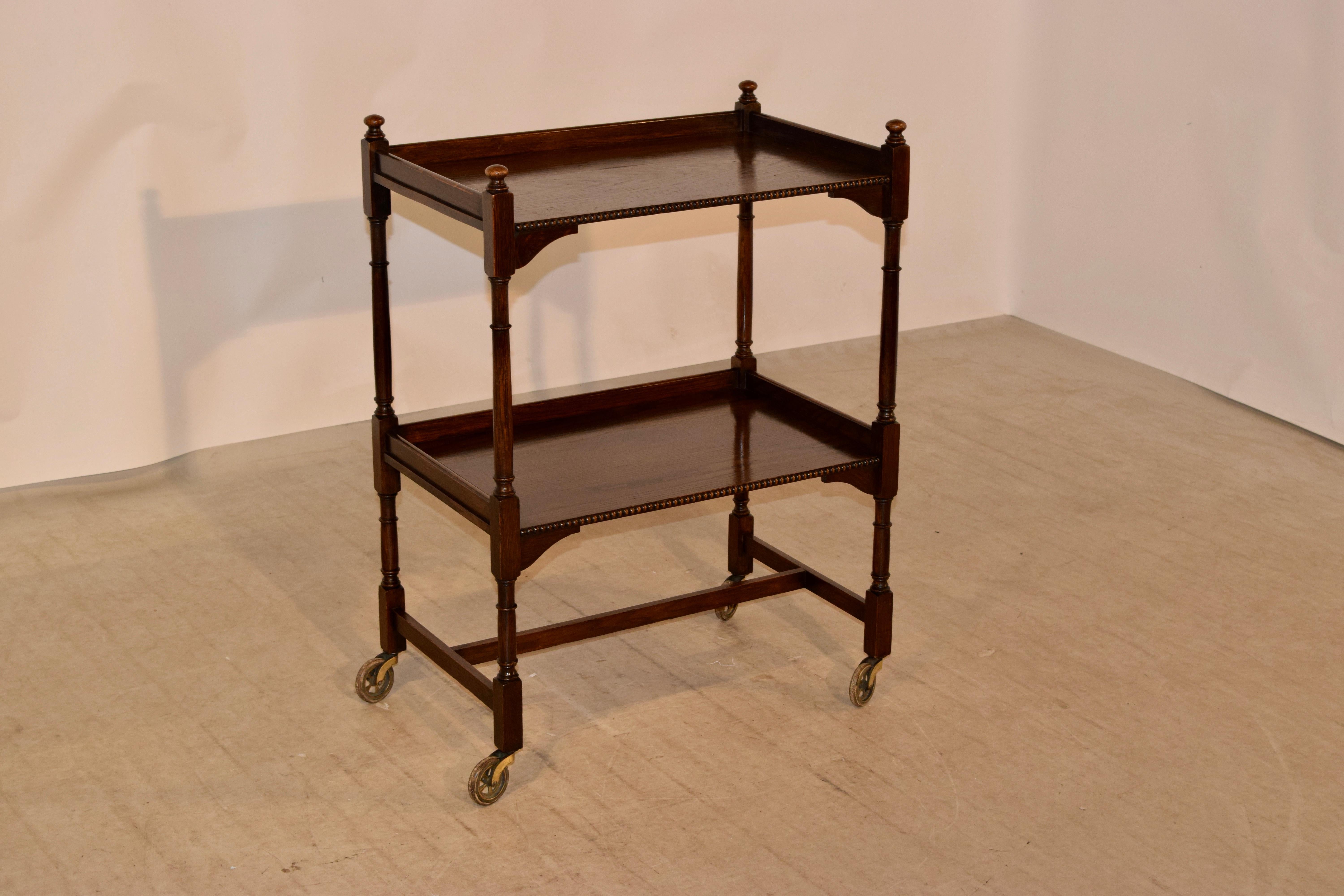 Oak drink cart from England with finials at the top over two shelves with beaded front edges, both with surrounding galleries and separated by hand-turned shelf supports. Raised on hand-turned legs joined by stretchers and brass casters.