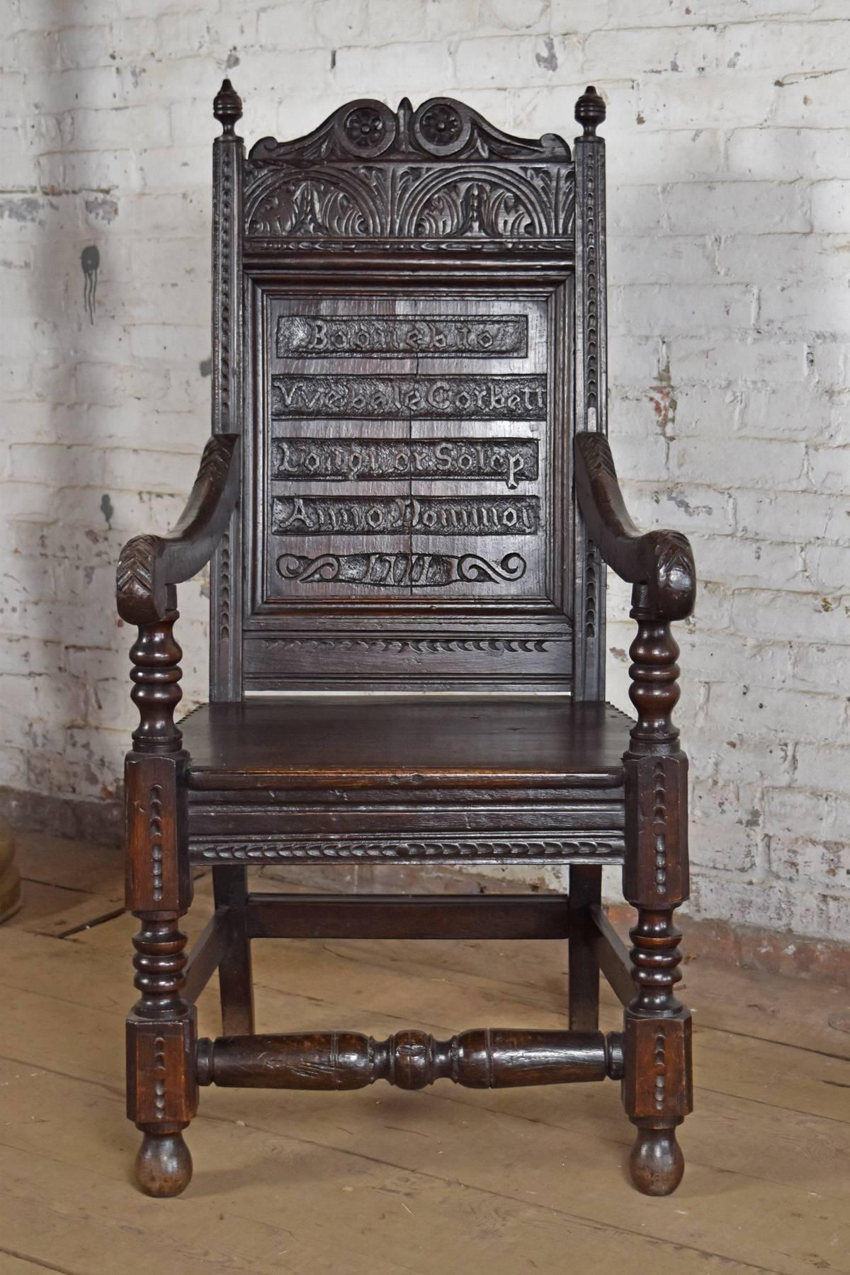 Open Armchair of typical Wainscot form, the straight paneled back adorned with carved lettering probably documenting family event, wavy armrests with Primitive carved decoration, paneled seat above a frieze, front legs and stretcher turned and chip