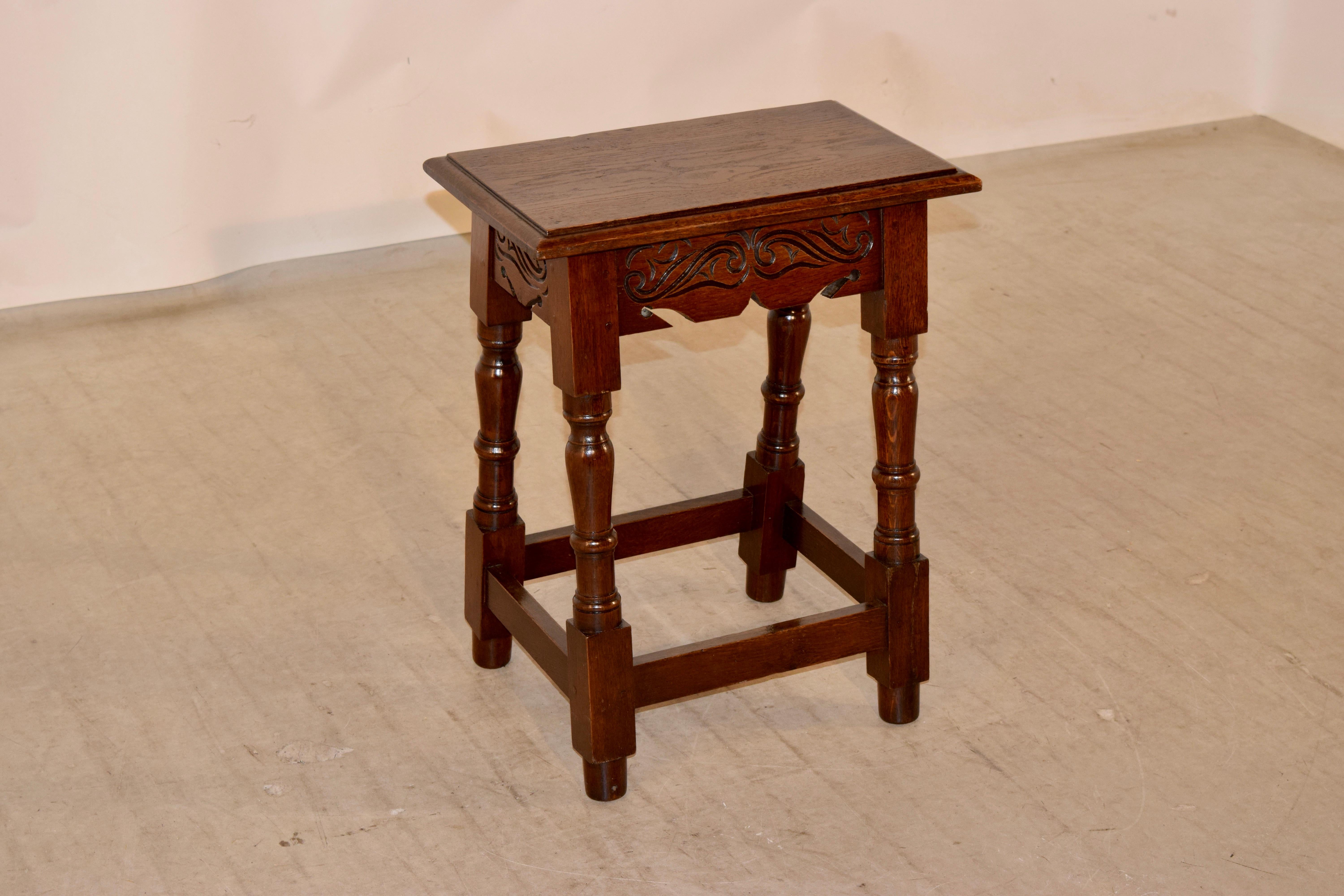 English oak joint stool with a beveled edge around the top, following down to a scalloped and hand carved decorated apron, over turned and splayed legs, joined by simple stretchers, circa 1920.