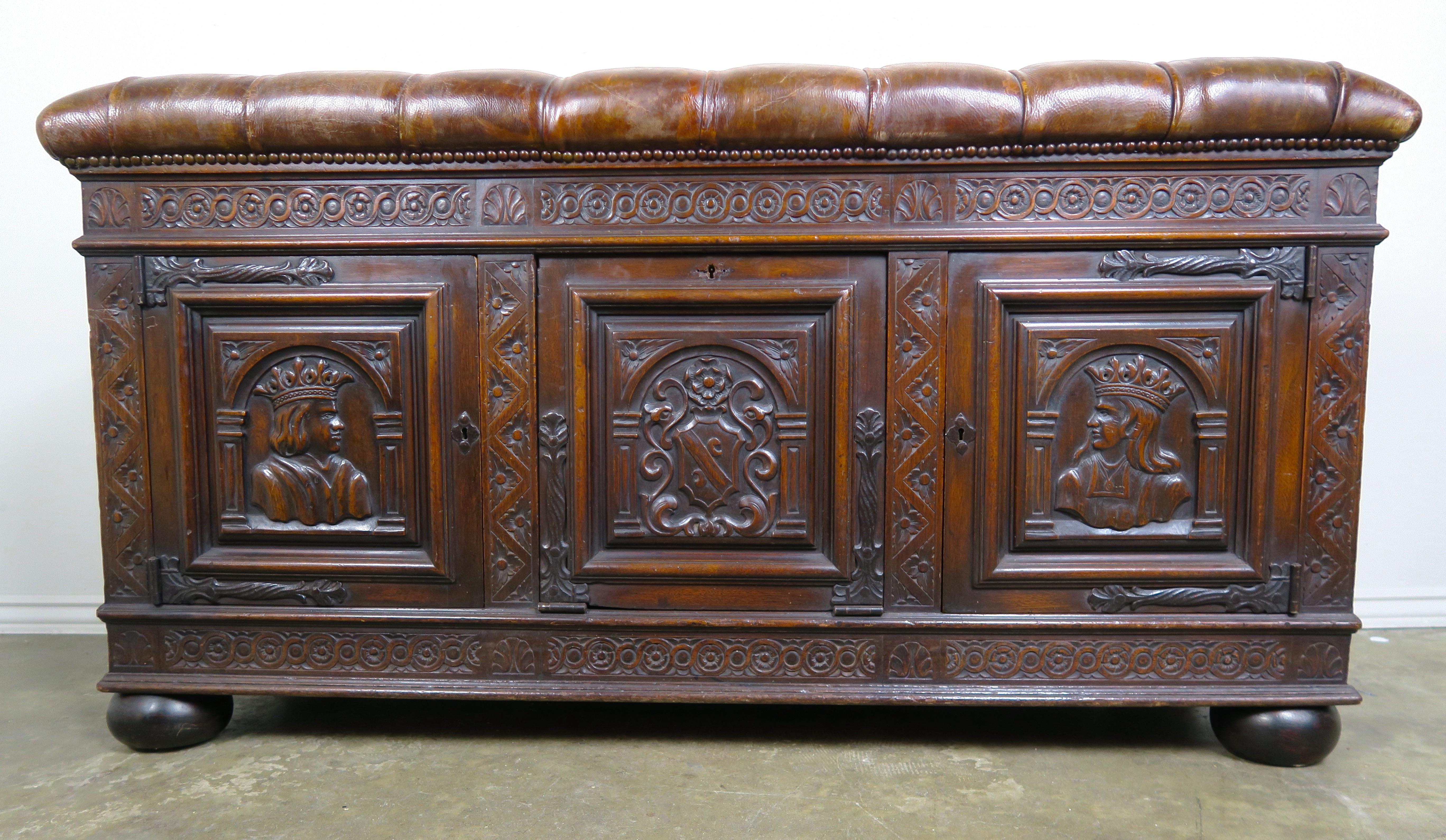English oak carved bench/trunk that is detailed with a coat of arms flanked by the king & queen. The top is upholstered in tufted leather and detailed with antique brass nail heads. The piece stands on four bun feet and has three doors that open to