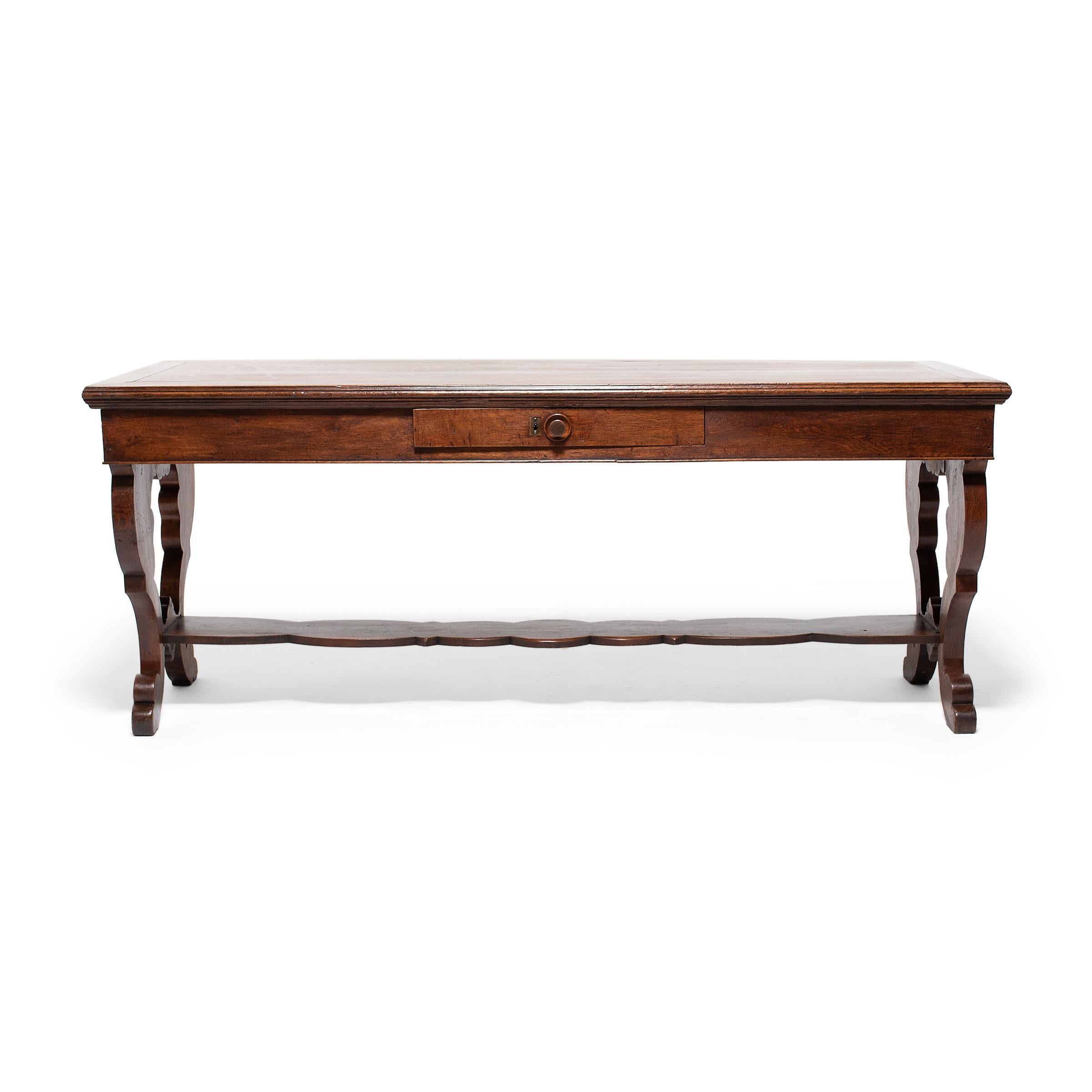 This grand oak writing table unites the refined flourishes of Regency-era English furniture with the timeless appeal of country antiques. Dated to the mid-19th century, the desk has a broad, rectangular top resting on stylized lyre-form end