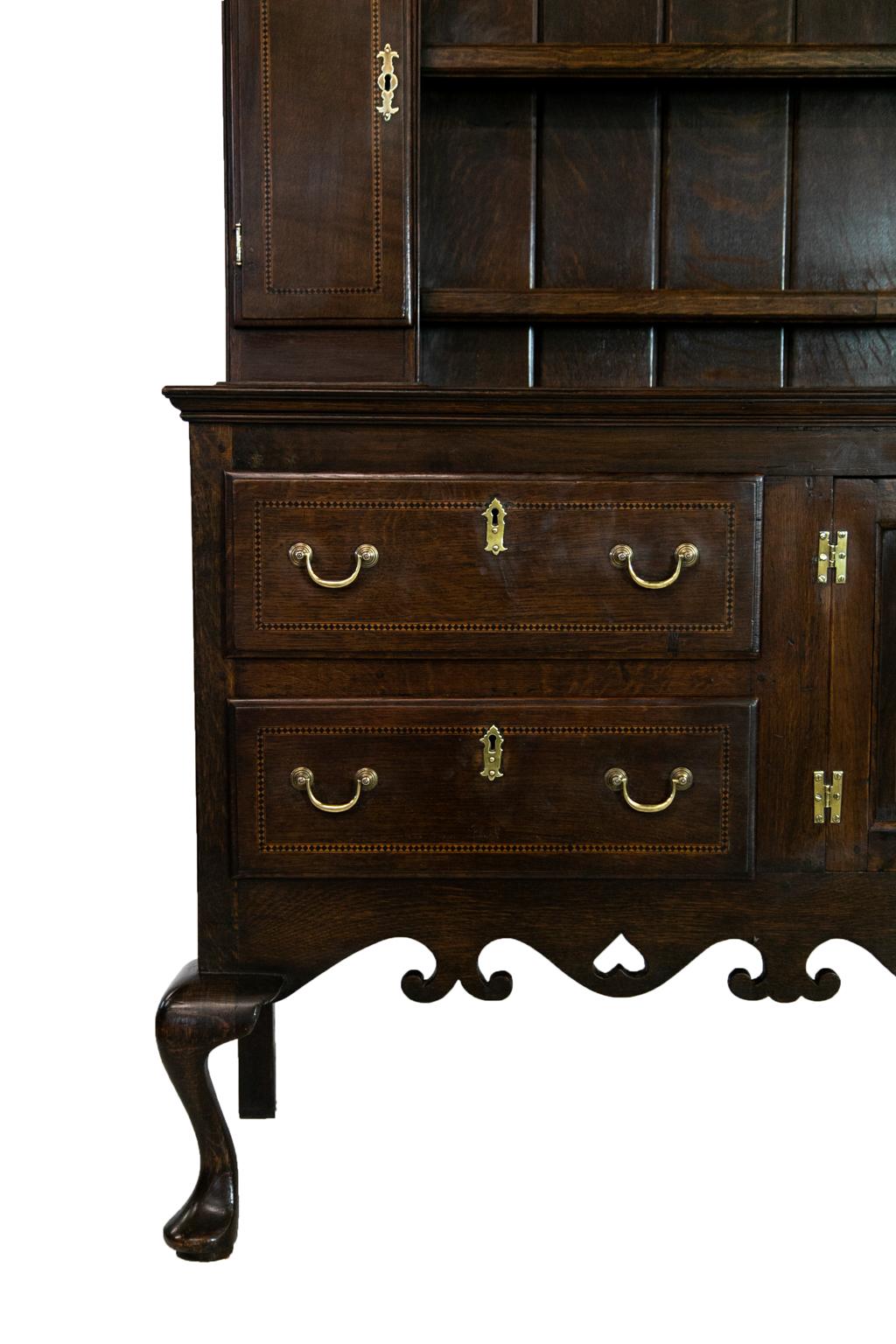 The cornice of this English oak Welsh dresser has a pierced apron. The top shelf is flanked by two arched openings that have scalloped shelves. The lower shelves of the top are flanked by inlaid cupboard doors. The lower section has two inlaid