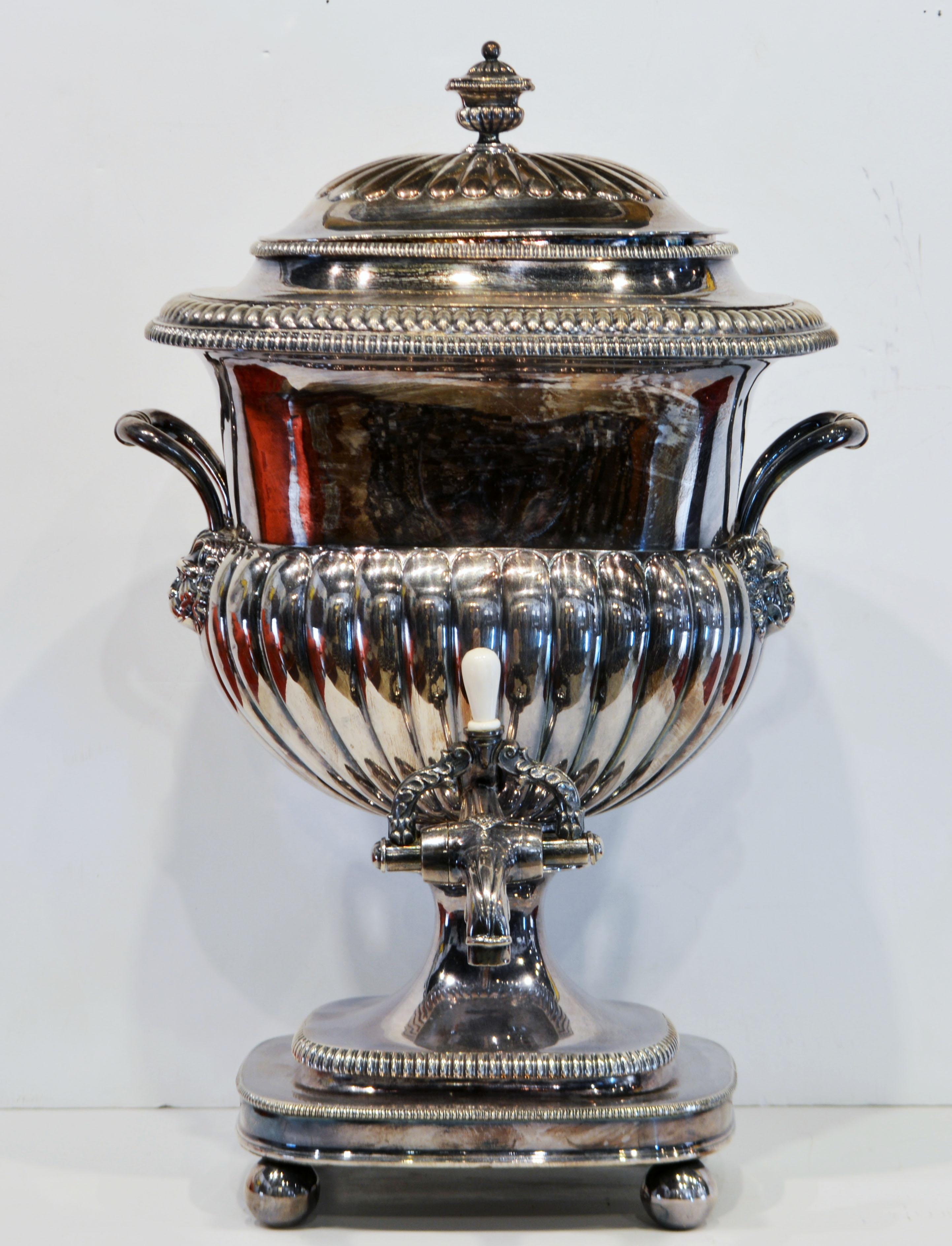 This regency Old Sheffield silver on copper tea urn features a half fluted lid with urn shaped finial, handles adorned with lion heads, polished bone tap handle, gadroon borders and a half fluted body and gadroon borders resting on four ball