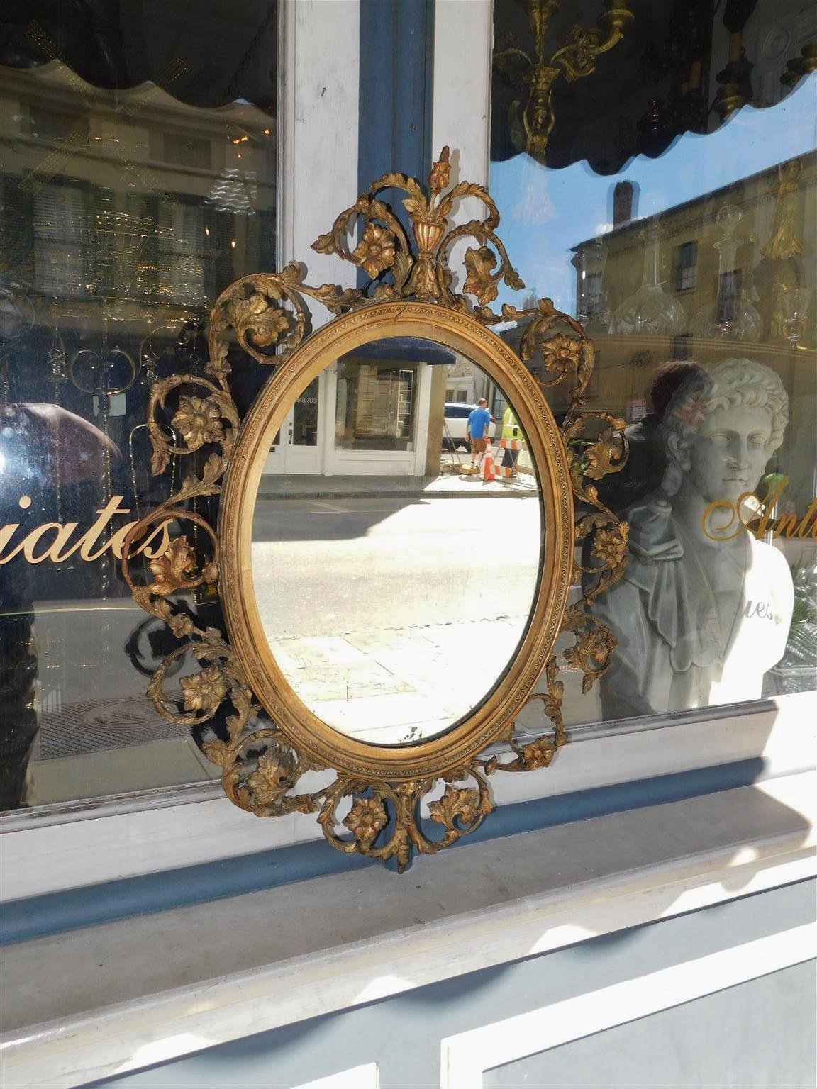 English oval gilt carved wood and gesso scrolled foliage wall mirror with centered floral urn crest. Early 19th century.