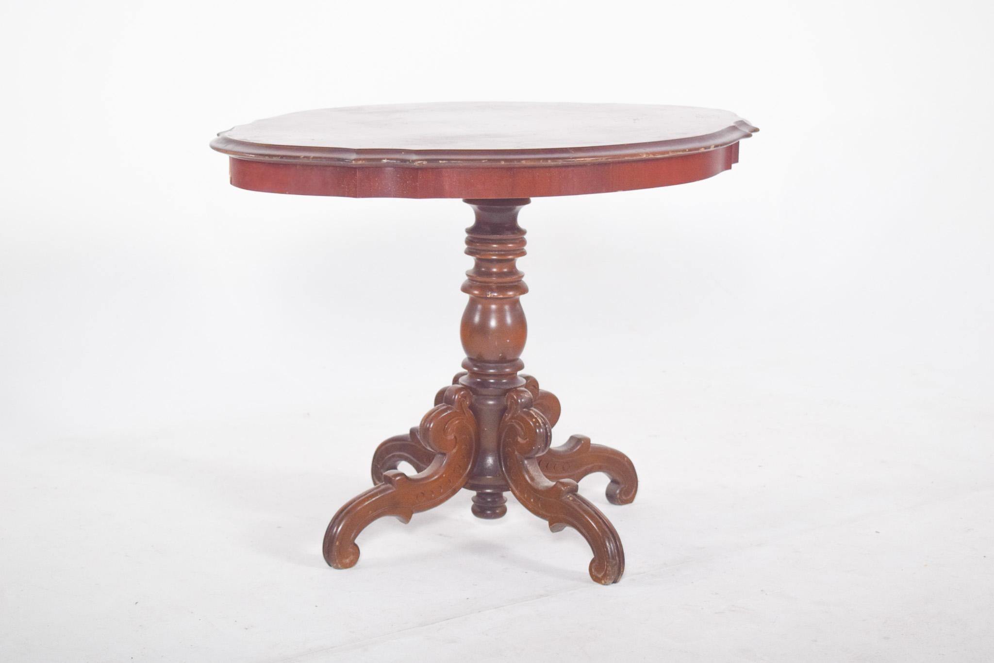 Oval coffee table. It rests on a turned central foot, based on four curved and carved supports, typical of Victorian inspiration. The frame is sturdy and well balanced, made of solid wood, probably mahogany or brown, with a varnished finish. The top