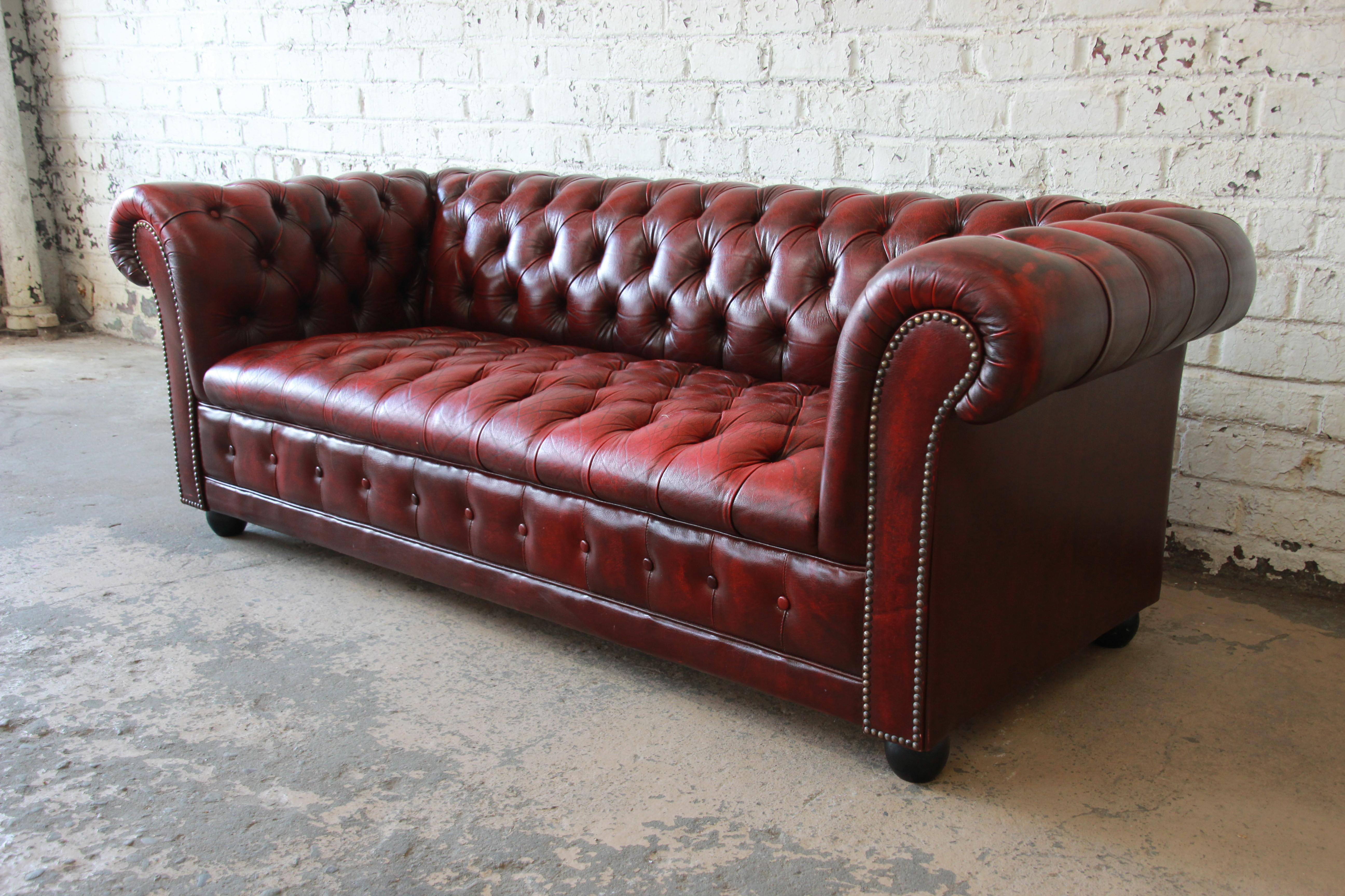 A gorgeous vintage English tufted oxblood red leather Chesterfield style sofa. The sofa features soft and extremely comfortable high grade tufted leather in oxblood red. The scrolled arms are brass studded, and the sofa sits on wooden bun feet. The