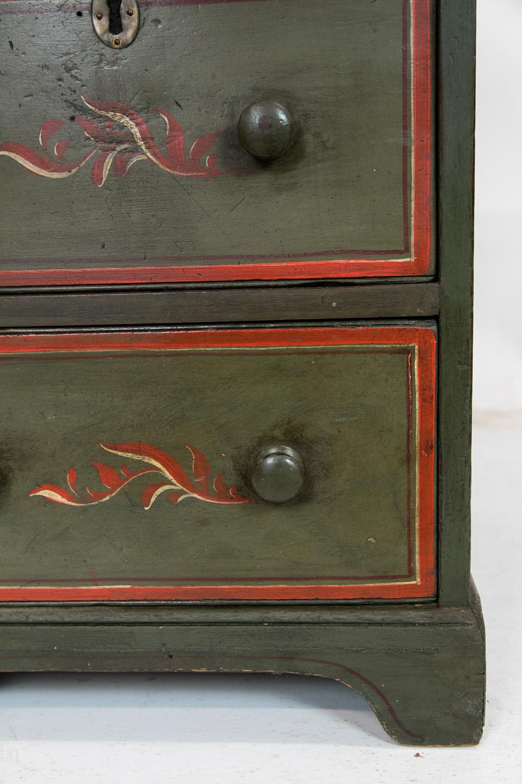 This chest has three drawers that are painted with red and white leaf designs. The top, drawer fronts, and sides are painted with red banding and the sides have recessed panels. The center drawer has an oval brass escutcheon. The inside lock mortise