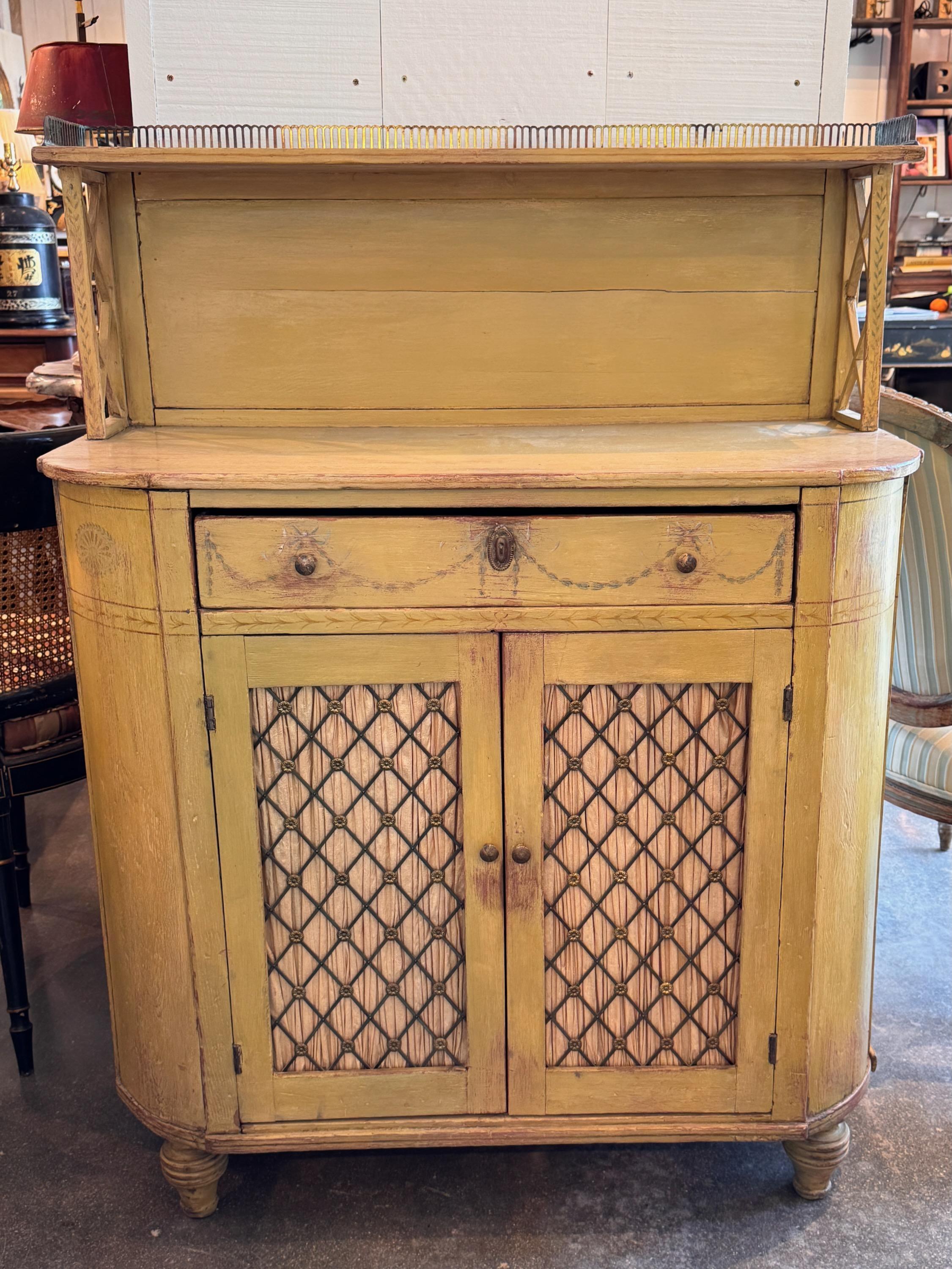 English painted chiffonier with a shaped top and pierced metal gallery, supported by open latticework sides. The drawer features painted swag detailing, and the lower cabinet has mesh-front doors backed in gathered fabric. Turned feet and natural
