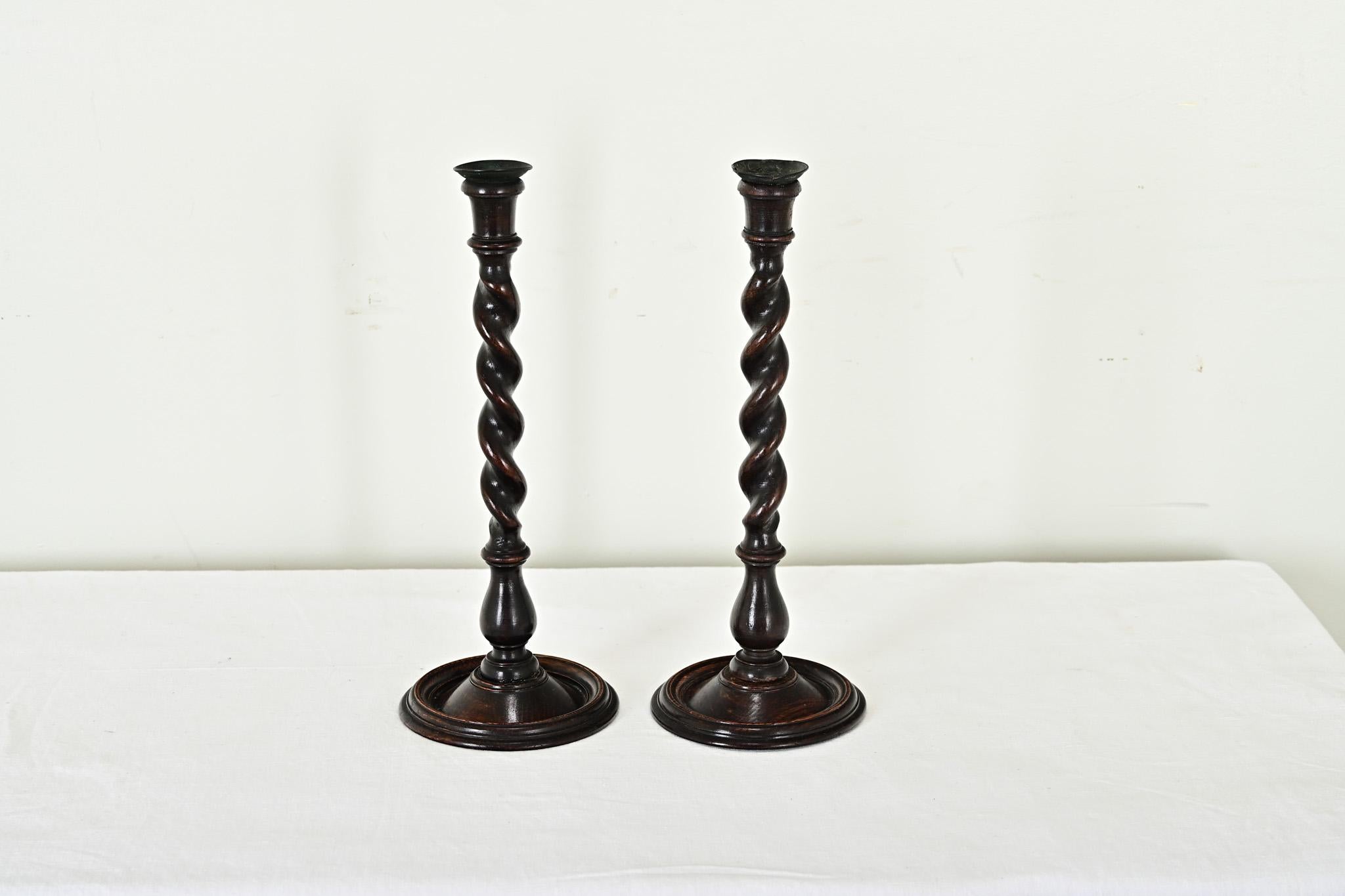 Elegantly carved pair of English oak barley twist candlesticks to grace your fireplace mantel or dining table. Each candlestick is topped with a metal bobeche for protecting the dark wood from hot wax. The twisting solid oak columns terminate as a