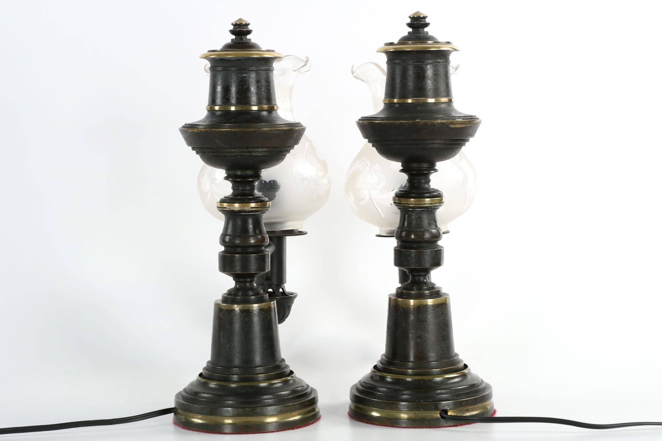 On this fine pair of electrified antique table lamps, the brass is patinated to a deep oiled black which is highlighted with a recurring strata of applied natural brass banding encircling the lamps. At the base, each lamp starts out with a brief