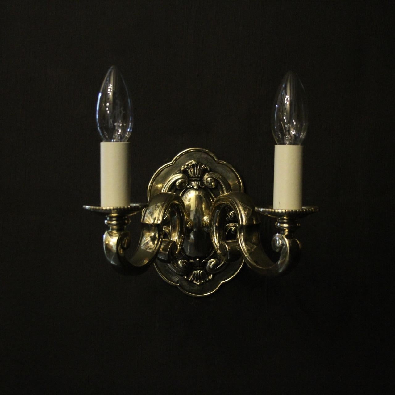 An English pair of repolished cast brass twin arm antique wall lights, the large gauge scrolling arms with circular bobeche drip pans, issuing from an ornate floral edged backplate, sympathecially repolished and great proportions. Fully rewired and