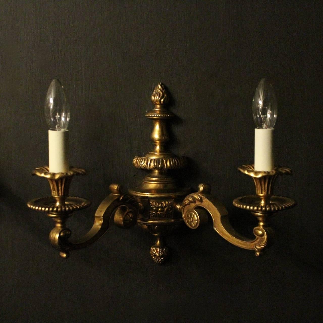 An English pair of gilded cast bronze twin arm antique wall lights, the square gauge leaf scrolling arms with circular leaf bobeche drip pans and trumpet reeded candle sconces, issuing from a decoratively cast elongated tapering backplate with flame