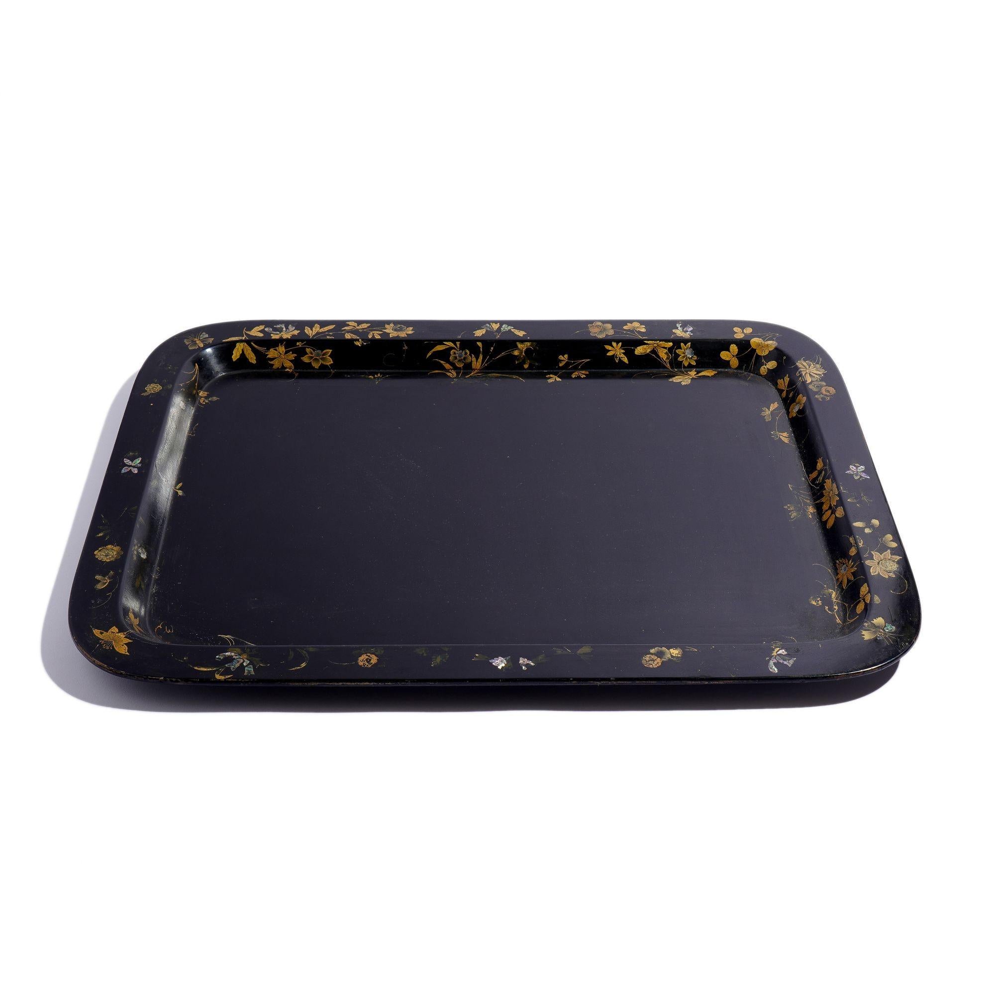 Large black flat rim paper mache tea tray. The rim of the tray is decorated with leafy gilt tracery and inlaid with abalone in the form of blooming periwinkles.
England, circa 1830.
