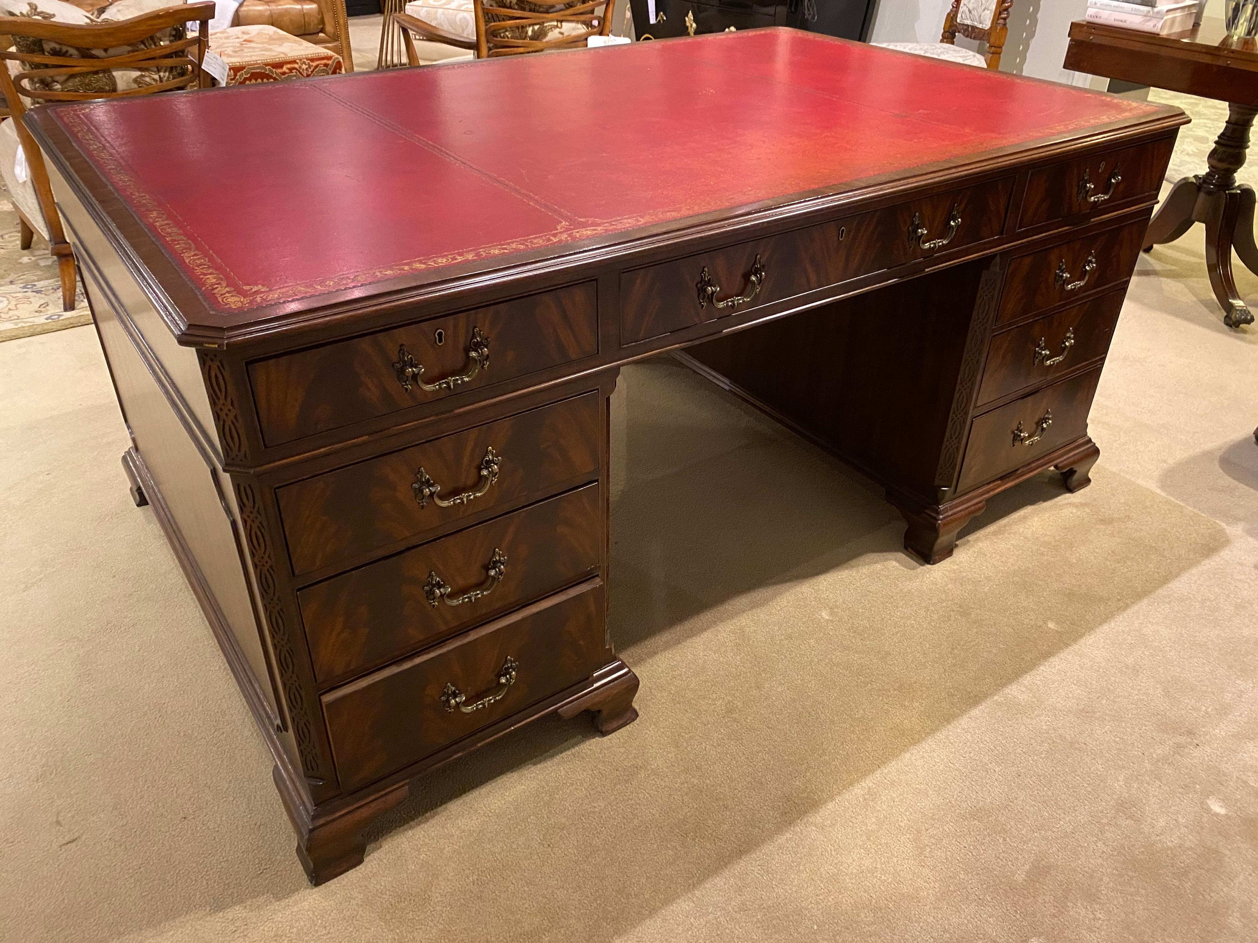 A classic English mahogany partners desk in Georgian style, with a red leather skiver top showing the character that comes with time. There is antique gold tooling around the edges. The desk has nine drawers on the executive side and doors on the