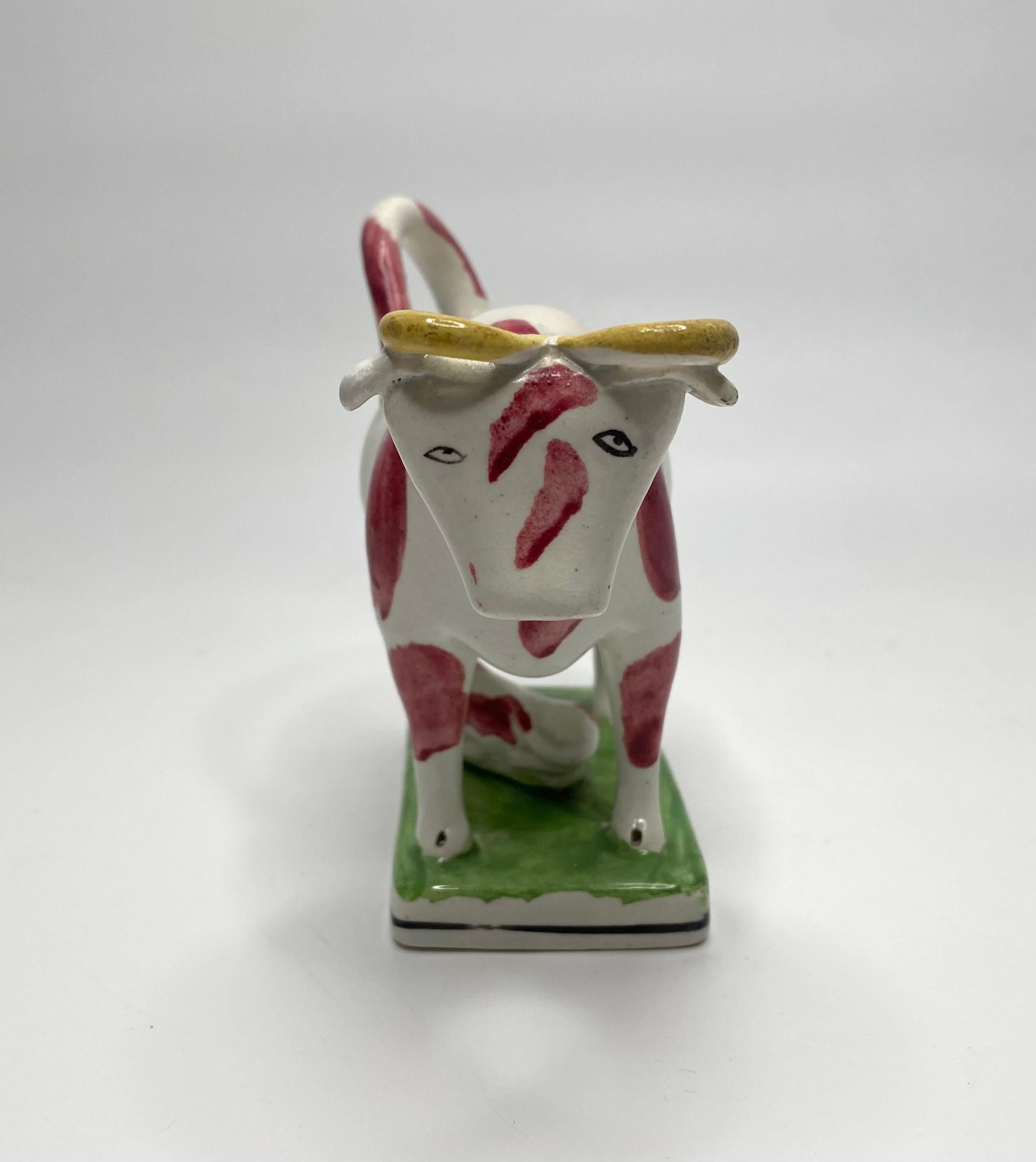 English pearlware cow creamer and cover, c. 1830. The Staffordshire or North England pottery cow, set upon a rectangular base, and painted with large puce spots, its horns yellow. The cow is accompanied by her calf, laid beneath her, on the grass