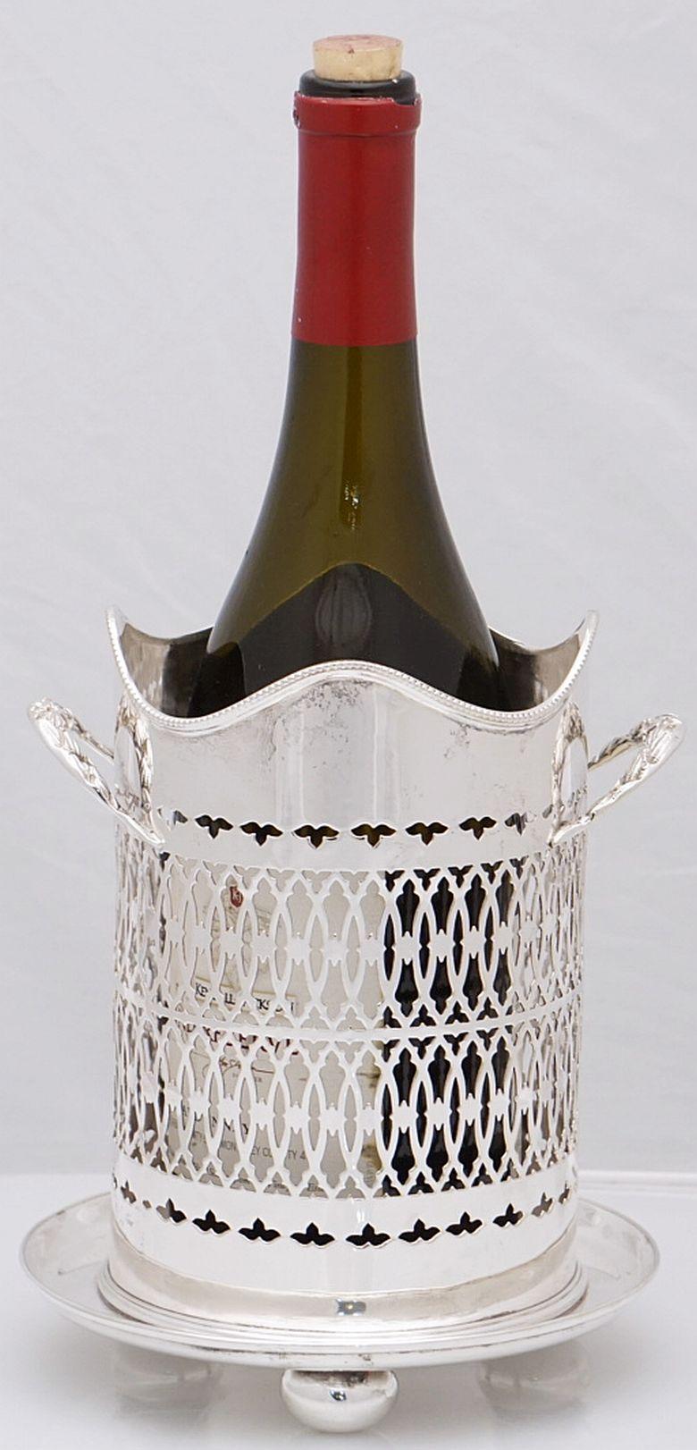 A handsome English wine bottle display holder or coaster of fine plate silver, featuring a rolled serpentine edge around the top, with an elegant pierced design around the circumference and opposing handles, and raised cylindrical base with ball