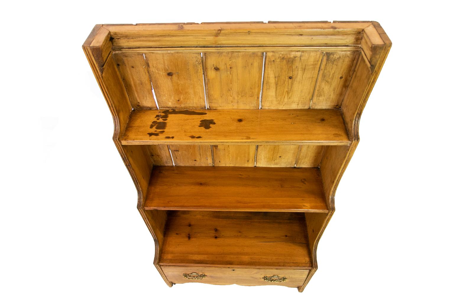 This pine book shelf has a heavy shaped molding supporting the top and has reeded front shaped sides framing three graduated shelves, which also have triple reeded front edges. There is a deep drawer below the bottom shelf and a shaped apron below