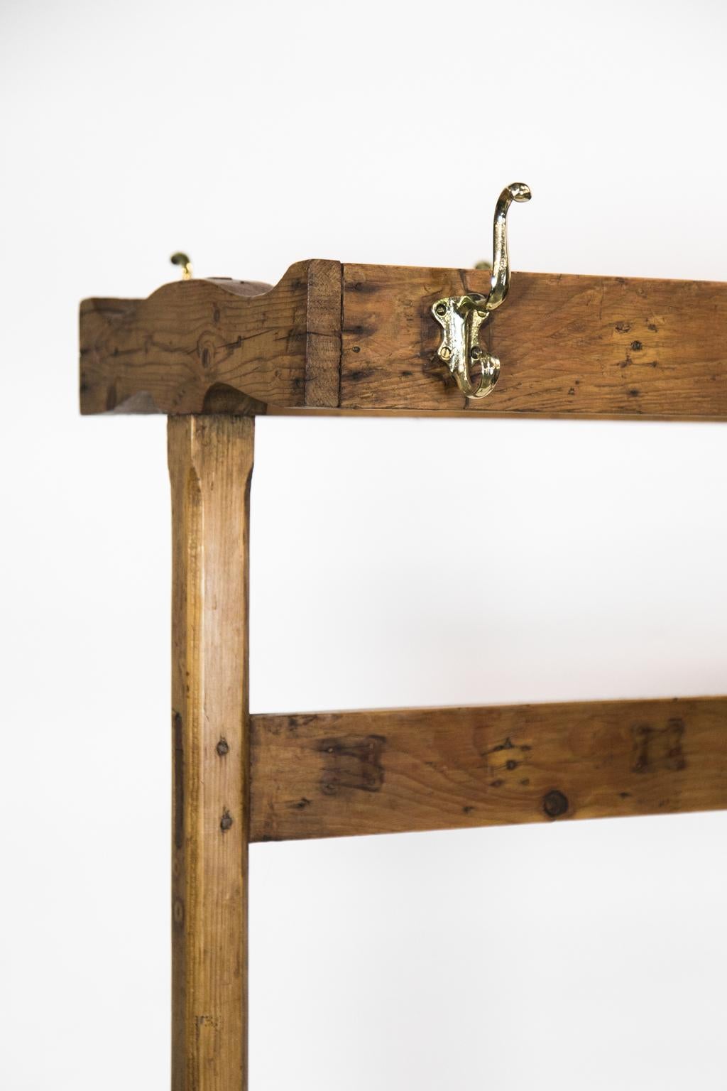 This pine rack has five heavy cast brass hooks on either side. There is double peg and exposed mortise and tenon construction. The side supports have chamfered pencil post corners. The bottom has a cross stretcher, and the legs terminate in a