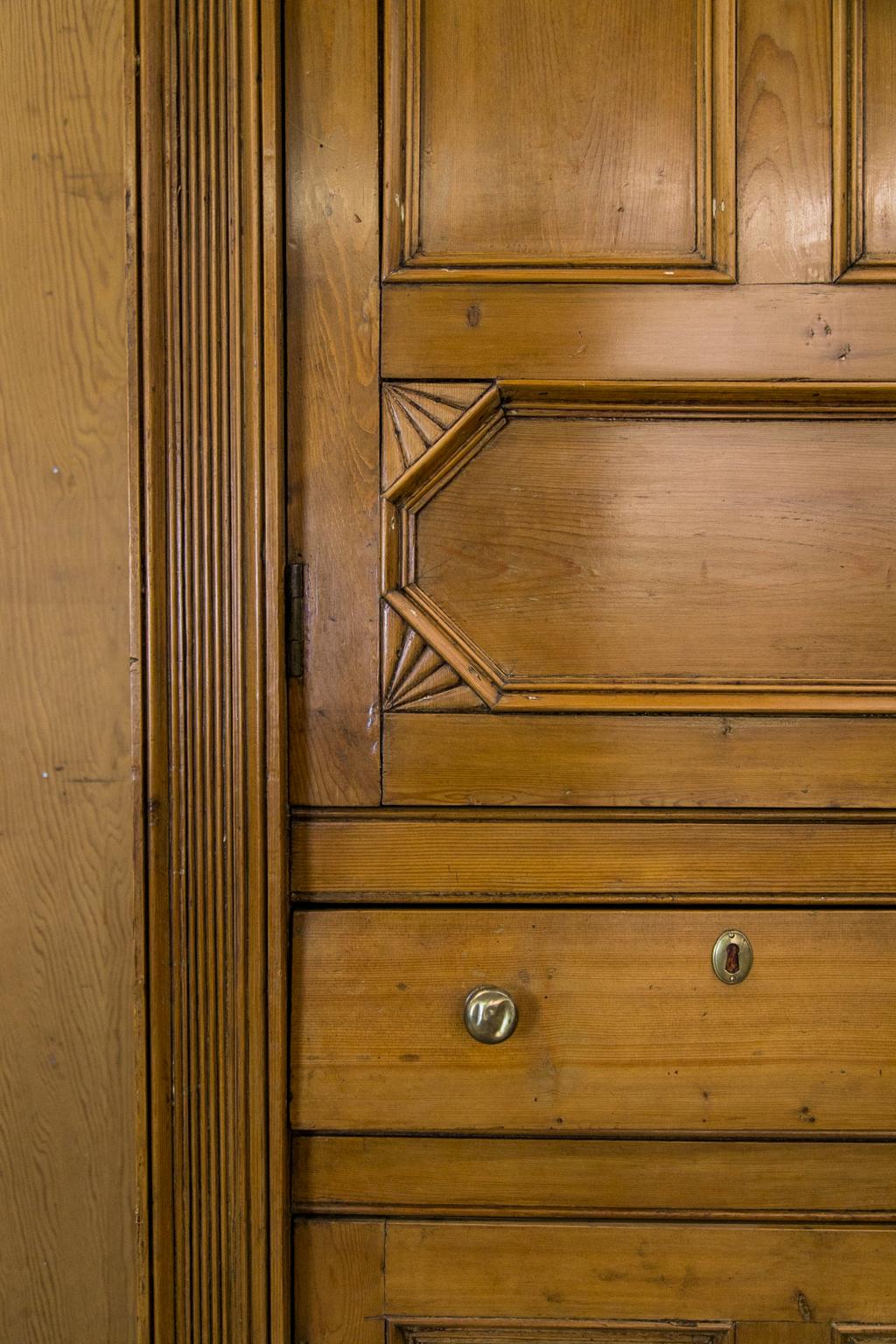 Cette armoire en pin anglais comporte deux portes supérieures avec deux panneaux en retrait encadrés de moulures sculptées et des panneaux inférieurs encadrés de quarts d'éventail stylisés sculptés. La corniche présente une frise sculptée et