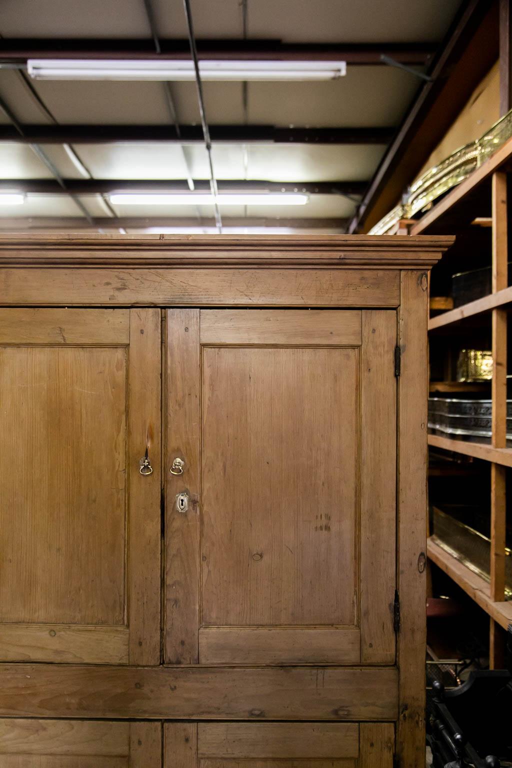 The upper section of this pine cupboard has one fixed shelf while the lower section is open. There is exposed peg construction. The doors have recessed panels with carved shaped molded edges. The brass hardware is later.