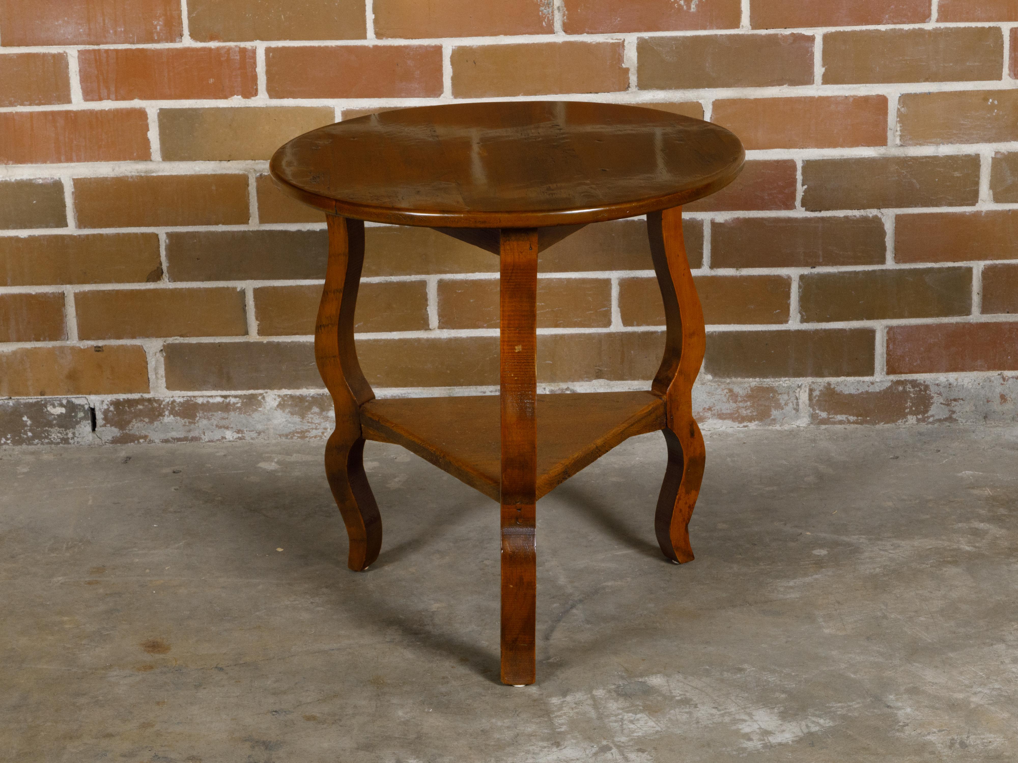 An English pine side table with circular top, curving legs and triangular shelf. This English pine side table, dating from the late 19th to early 20th century, offers a blend of rustic charm and elegant design with its circular top and trio of