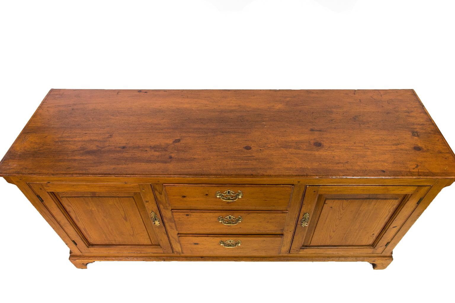 Dieses englische Sideboard aus Kiefernholz hat eine freiliegende Zapfenkonstruktion. Die drei Schubladen haben umgeschlagene Kanten. Die Türen sind mit wulstigen Zierleisten umrahmt und haben erhabene Füllungen. Die Ecken sind abgeschrägt. Der