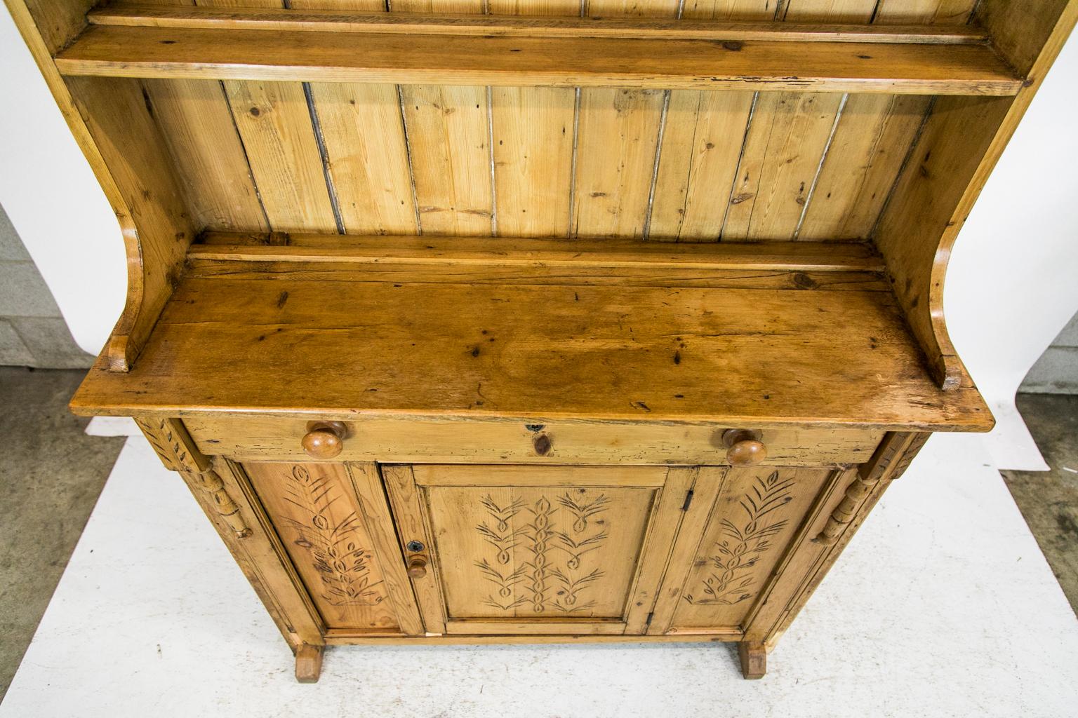 This pine cupboard has two shelves and a serving platform that all have an attached plate rail. The crown has a heavy dentil molding and the lower door and side panels have incised carvings of stylized leaf and flower designs. The wooden knobs and